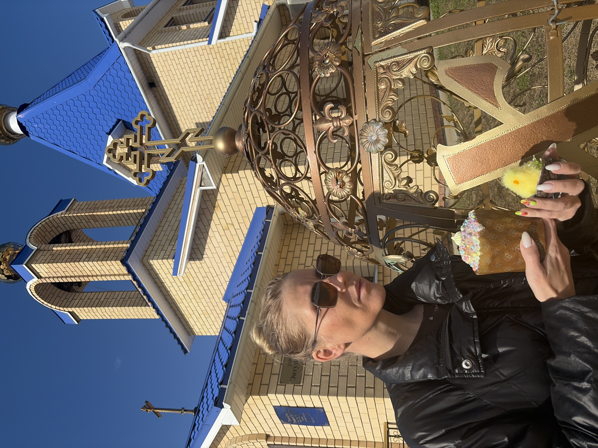 A woman in a black jacket and sunglasses holds an Easter cake and a chick toy in front of a golden metal gate and the Church of the Holy New Martyrs and Confessors of Russia in Chaikovsky, Russia. The church is constructed of light-colored bricks with blue roofs and trim. The gate has a large Cyrillic letter "X" on it. The sky is blue and cloudless.