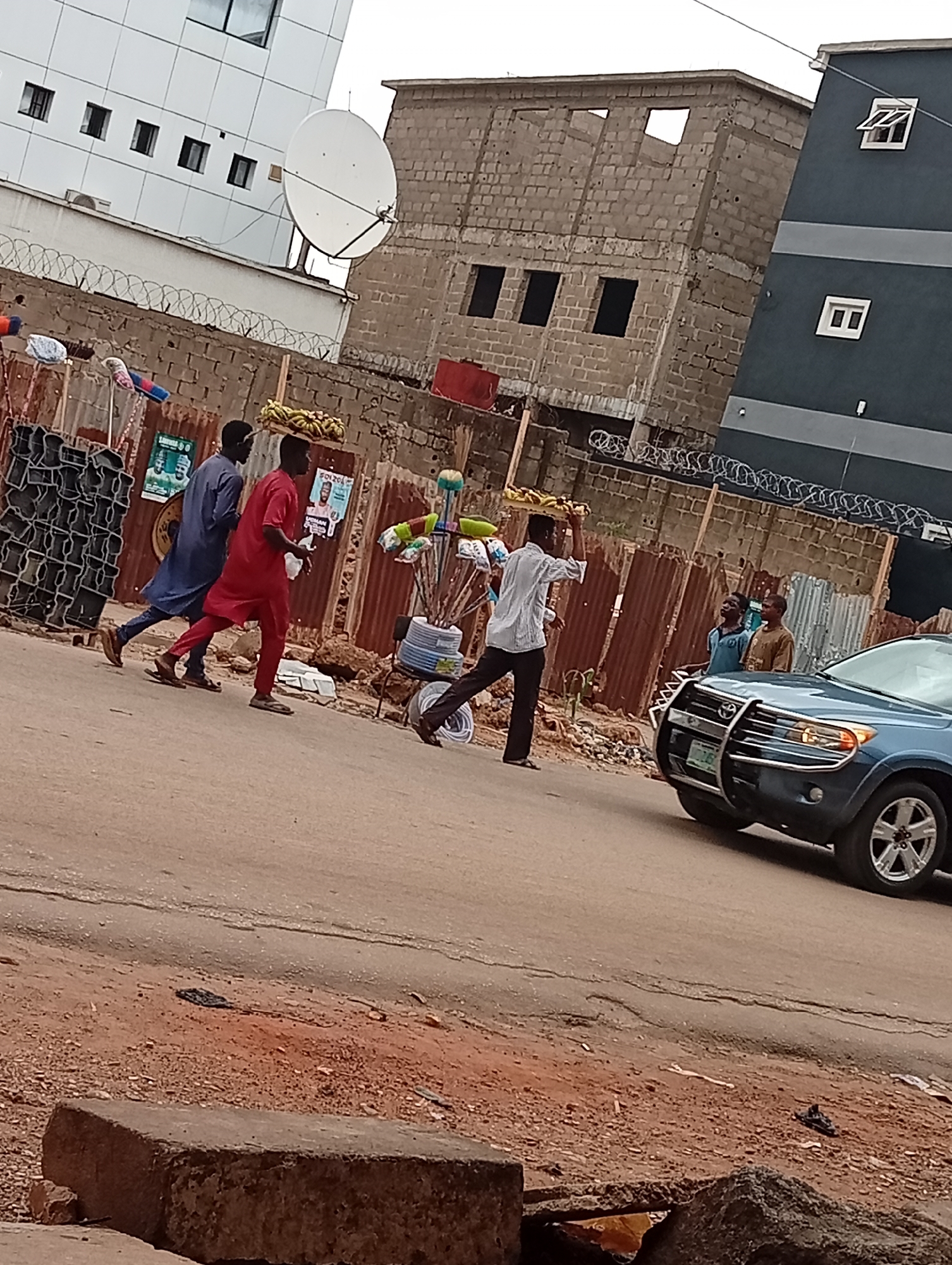 The image depicts a street scene in Maiburiji, Nigeria. Three men carry goods on their heads, walking along a road. The first man wears a blue robe, the second a red one, and the third a light-colored shirt and dark pants. A blue SUV is driving on the road. Behind the men are buildings, including one under construction. The street is paved but shows signs of wear and tear. In the foreground is a weathered stone or concrete block. A satellite dish is visible on one of the buildings. Barbed wire tops a fence running along the side of the road.
