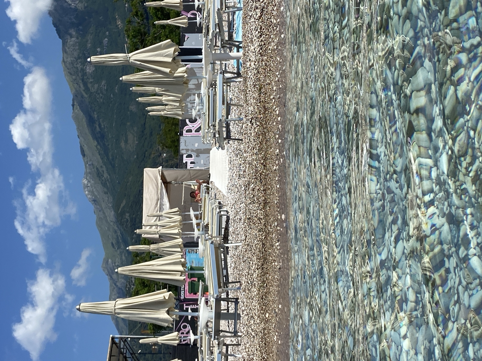 The image depicts a sunny day at a pebbly beach in Budva, Montenegro, viewed from within the clear, shallow water. In the immediate foreground, transparent water reveals a bed of various-sized light-colored pebbles and rocks, with light ripples reflecting on the surface. The midground features a wide, light brown pebble beach. Arranged across the beach are multiple rows of closed, cream-colored beach umbrellas, accompanied by empty light-colored lounge chairs and small tables. Towards the left midground, a dark sign with the pink letters "PRO" is visible, along with parts of metal scaffolding. In the center, a light-colored tent structure stands near green refuse bins and another partially visible sign reading "GOSTL TE". On the far right, another large, light-colored tent houses a single person in a red garment, reclining on a lounge chair. The background is dominated by large, green, rugged mountains under a bright blue sky dotted with scattered white cumulus clouds.