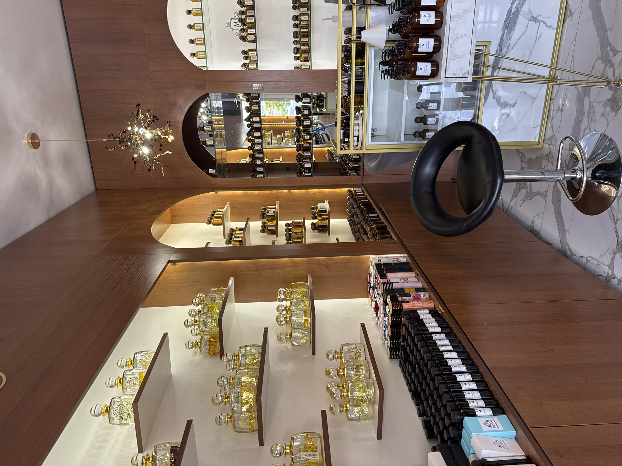 This media file depicts the interior of a perfume shop in Krasnodar, Russia. The scene is dominated by wooden shelves and displays showcasing a wide variety of perfumes in ornate glass bottles. The lighting is warm and inviting, suggesting it is daytime, possibly late afternoon, as the interior lights are on.
The shelves are organized with multiple rows of perfume bottles, some appearing to be antique or luxury items, with gold accents and intricate designs. There are also displays of larger bottles, possibly diffusers or cologne, arranged on higher shelves. A prominent black bar stool with a chrome base is positioned in the foreground, near a tall, slender gold floor lamp. In the background, a marbled wall feature adds a touch of elegance.
The overall atmosphere is one of a high-end boutique, emphasizing the curated collection of fragrances. The arrangement of the products suggests a retail environment designed to showcase the perfumes attractively to potential customers. No people are visible in the frame, and the focus is entirely on the products and the shop's sophisticated interior design. There are no discernible weather cues as the image is entirely indoors. Some labels are visible on the bottles and boxes, but the text is too small to be legible.
