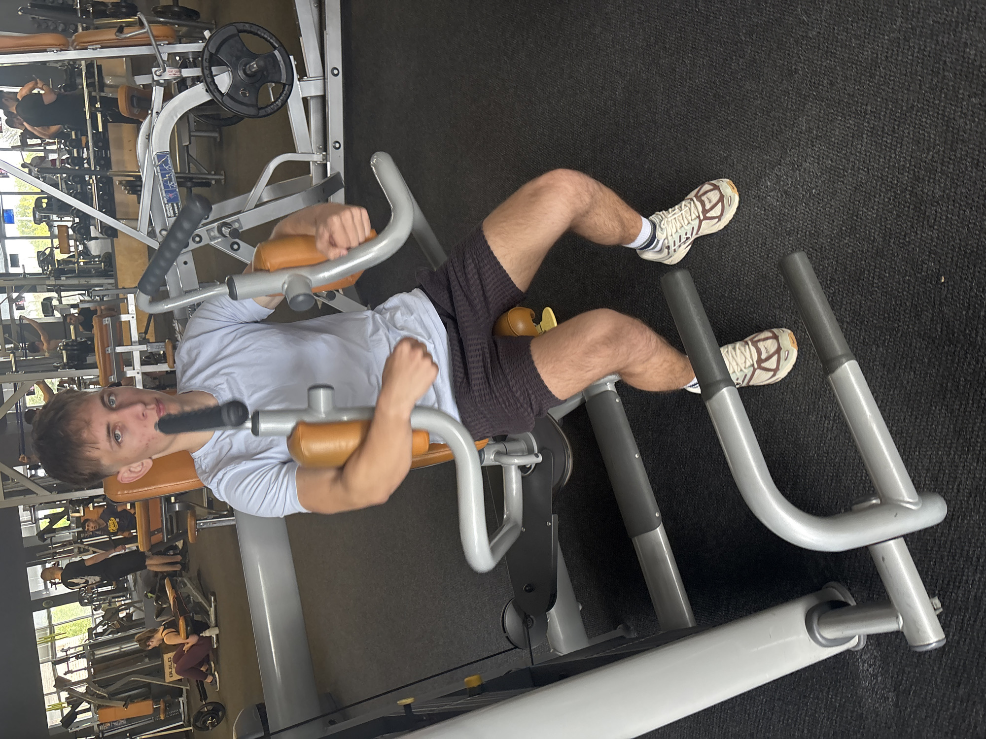 A young man is captured mid-workout in a gym located in Blagoevgrad, Bulgaria. He is seated on a hip abduction/adduction machine, positioned with his legs spread against the machine's pads. He wears a light blue t-shirt, dark grey athletic shorts, and light-colored sneakers with brown accents, paired with white socks. His hands grip the machine's handles, and he looks directly at the camera with a neutral expression. He has short brown hair and light stubble.
The background reveals a bustling gym environment with various other fitness equipment, including weight racks, benches, and several other machines. Other gym-goers are visible in the blurred background, engaged in their own exercises. Large windows suggest natural light is illuminating the gym, indicating it is daytime. The floor is covered with dark, textured gym matting. The text "BUILD" is faintly discernible on a piece of yellow and black equipment in the background. The image is taken from a slightly elevated perspective.