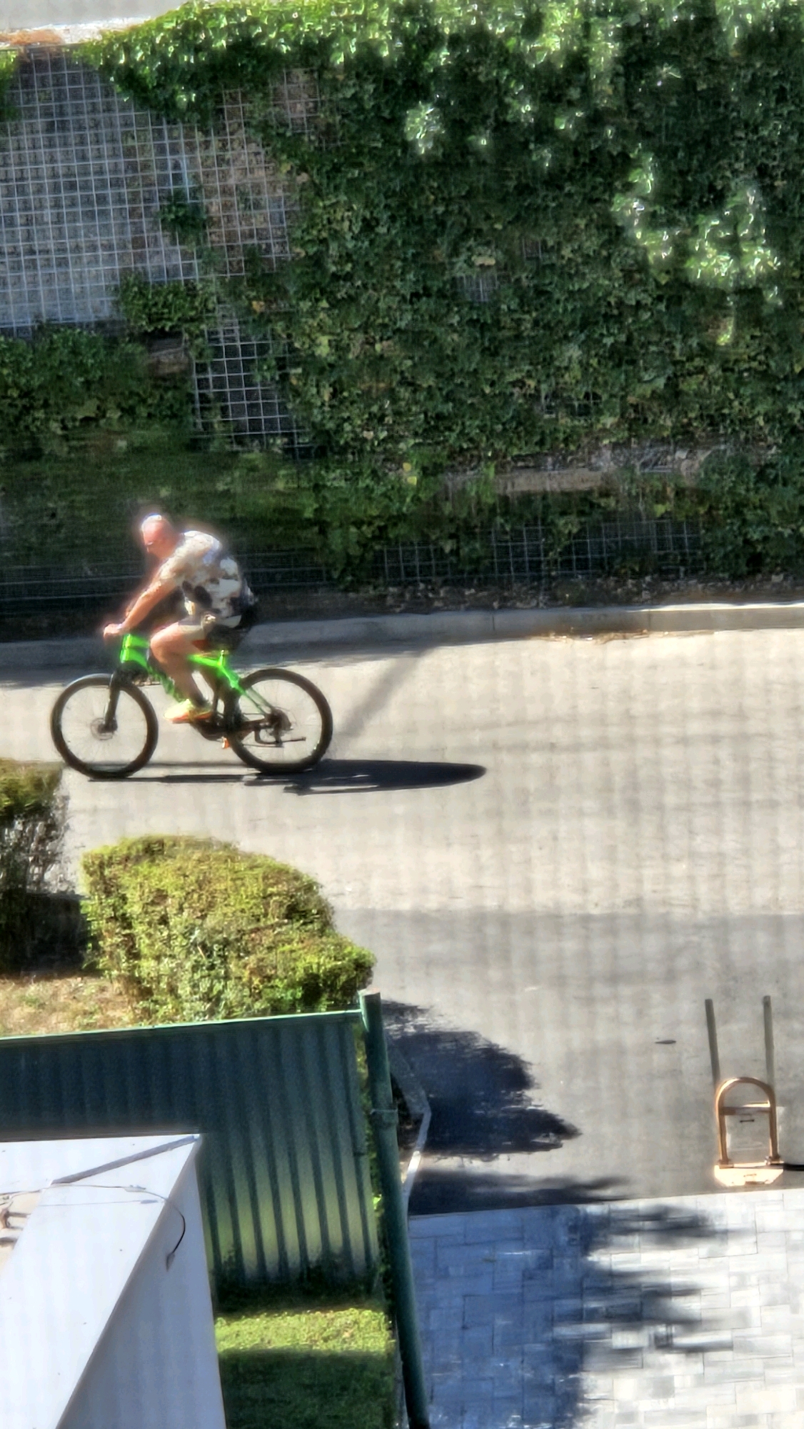 A person is riding a green bicycle on a paved road. The person is wearing a patterned shirt and shorts. The road is lined with vegetation on the left and a wall covered in ivy on the right. The sky is visible in the upper portion of the image, with sunlight filtering through the leaves of trees. The scene appears to be an outdoor environment, possibly a park or a residential area, in the city of Prešov, Slovakia.