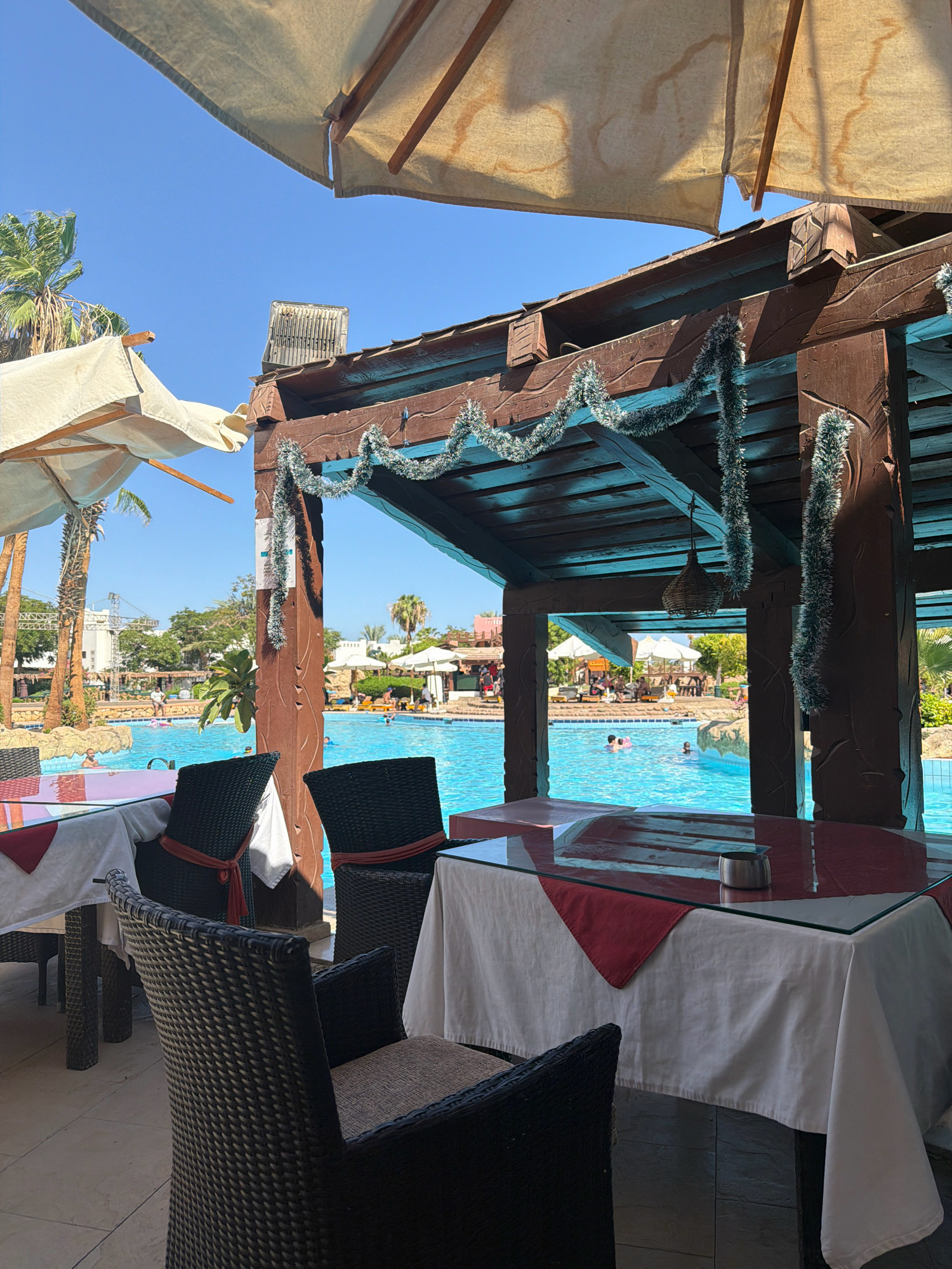 This is a photograph of an outdoor dining area adjacent to a swimming pool at a resort in Sharm El Sheikh, Egypt. The perspective is from under a covered structure with a wooden beam ceiling and decorative tinsel. In the foreground, there are tables set with white tablecloths and red accents, along with wicker chairs. Beyond the tables, a bright blue swimming pool is visible, with several people swimming and lounging on the side. Palm trees and resort buildings are present in the background, under a clear blue sky. The scene suggests a warm, sunny day at a vacation destination.