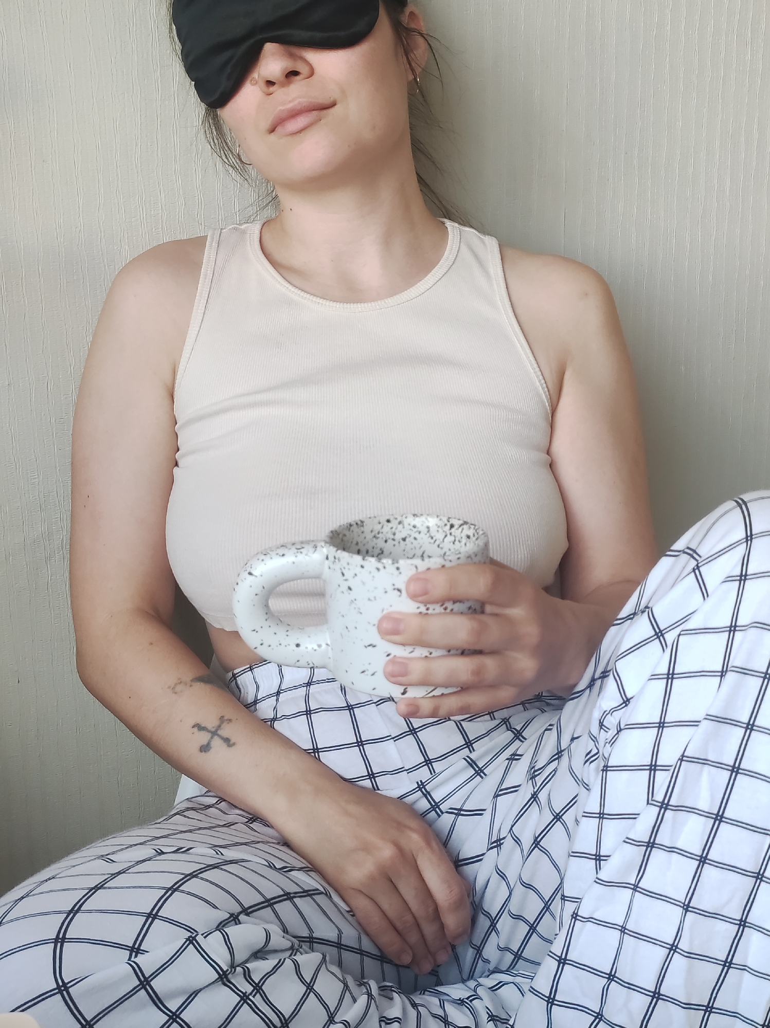 A woman is depicted sitting indoors, likely in a bedroom or living space in Chaikovsky, Russia. She wears a black sleep mask covering her eyes, suggesting a morning or pre-sleep moment. Her light-colored hair appears pulled back. She is dressed in comfortable sleepwear, consisting of a light beige ribbed tank top and white pajama bottoms featuring a distinct black grid pattern.
Both of her hands gently hold a white mug adorned with black speckles, implying she is either having a warm beverage or about to. Her facial expression, partially visible around the sleep mask, shows a soft, relaxed demeanor with a slight hint of a smile. On her left forearm, a small tattoo resembling two crossed bones or oars is visible. The background is a plain, light-colored wall with subtle vertical texturing. The overall scene conveys a sense of calm and leisure. There is no visible text in the image.