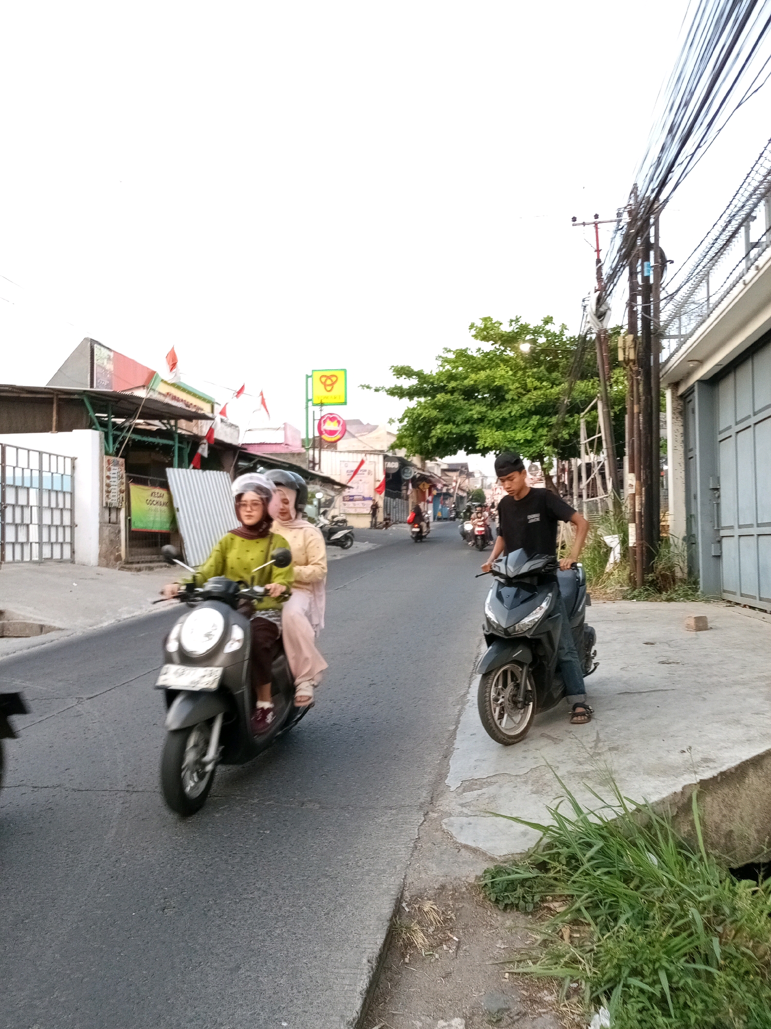 The image depicts a street scene in Sariwangi, Indonesia. In the foreground, two women wearing hijabs are riding a dark-colored scooter, with the driver in a lime green shirt and the passenger in a beige outfit. They are wearing helmets. Behind them, on the right side of the street, a young man wearing a black t-shirt and a black cap is seated on a black scooter. He is looking down and has his right hand on the handlebar. The street is paved and has some cracks. On the right side of the street, there is a concrete sidewalk with tall grass and a drainage ditch. Further down the street, other vehicles and buildings are visible. The buildings appear to be shops or residences, with some displaying signs and flags. Electrical wires are strung overhead, supported by poles. The sky is overcast and bright.
