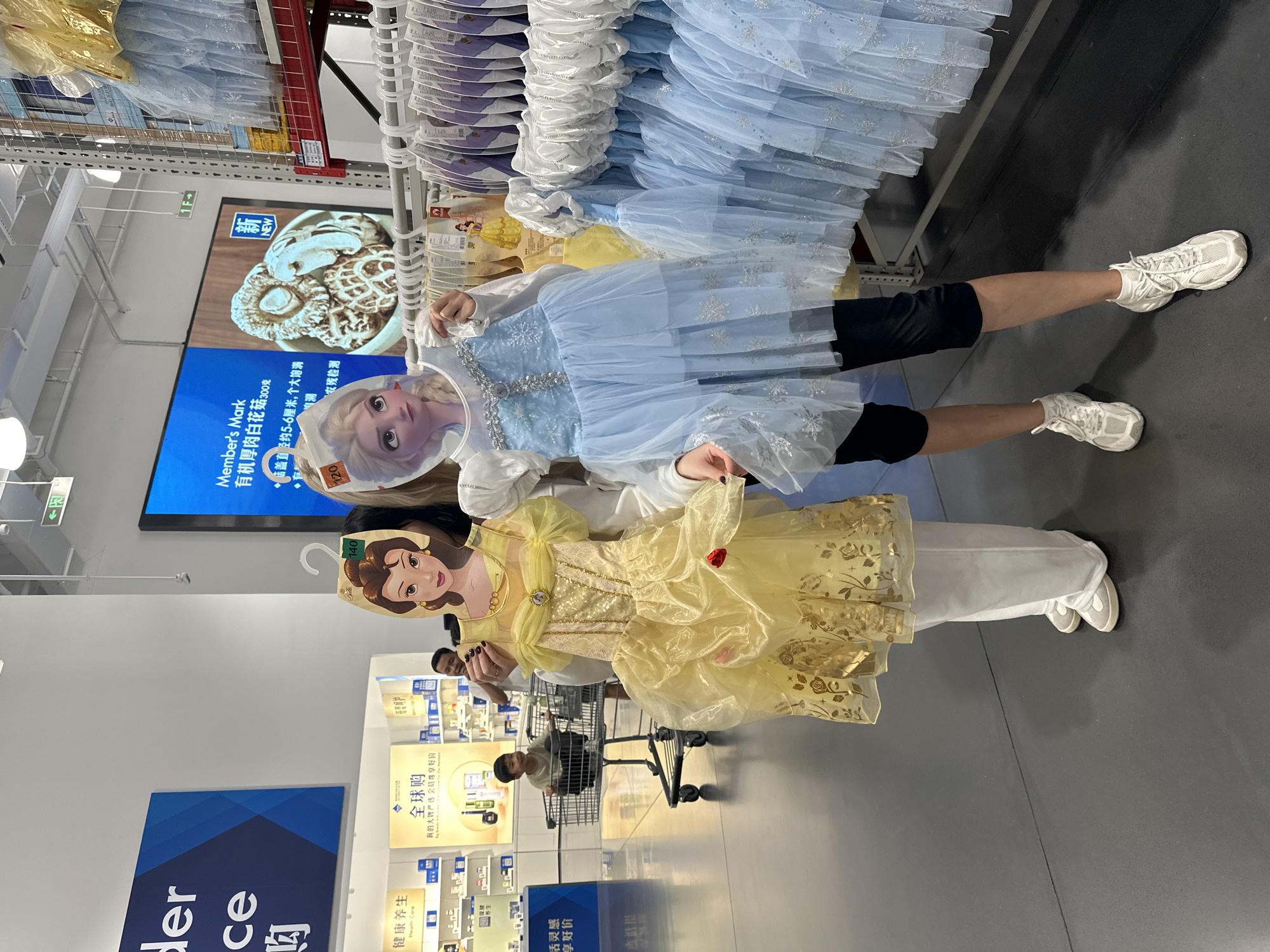 In a brightly lit retail store, possibly a wholesale club, two young women stand in an aisle displaying children's costumes. The woman on the left holds up a yellow Belle-inspired dress, complete with a character mask. She is wearing white pants and white sneakers. The woman on the right is dressed in a blue Elsa-inspired costume and black leggings, paired with white sneakers. Behind them, a young boy sits in a shopping cart, looking towards them. The background shows shelves stocked with merchandise, including more children's dresses, and overhead signs with Chinese characters. The store appears to be located in Liwan District, China, as indicated by context clues from similar images.