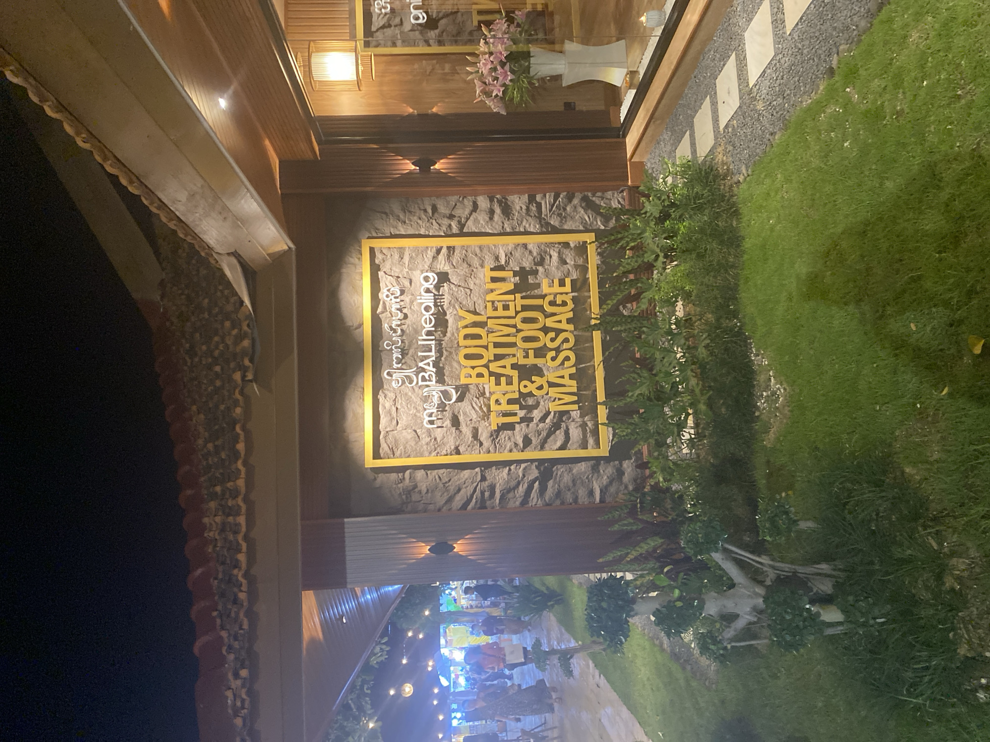 The image shows the exterior of a business establishment, possibly a spa or massage center, located in Kuta Selatan, Indonesia. The building's facade features a sign with the text "BALIhealing" and "BODY TREATMENT & FOOT MASSAGE" in yellow lettering. The sign is mounted on a stone-textured wall. The storefront includes a display window with plants. A walkway with people and string lights in the background leads further into the property. A strip of grass and landscaping are on the right.