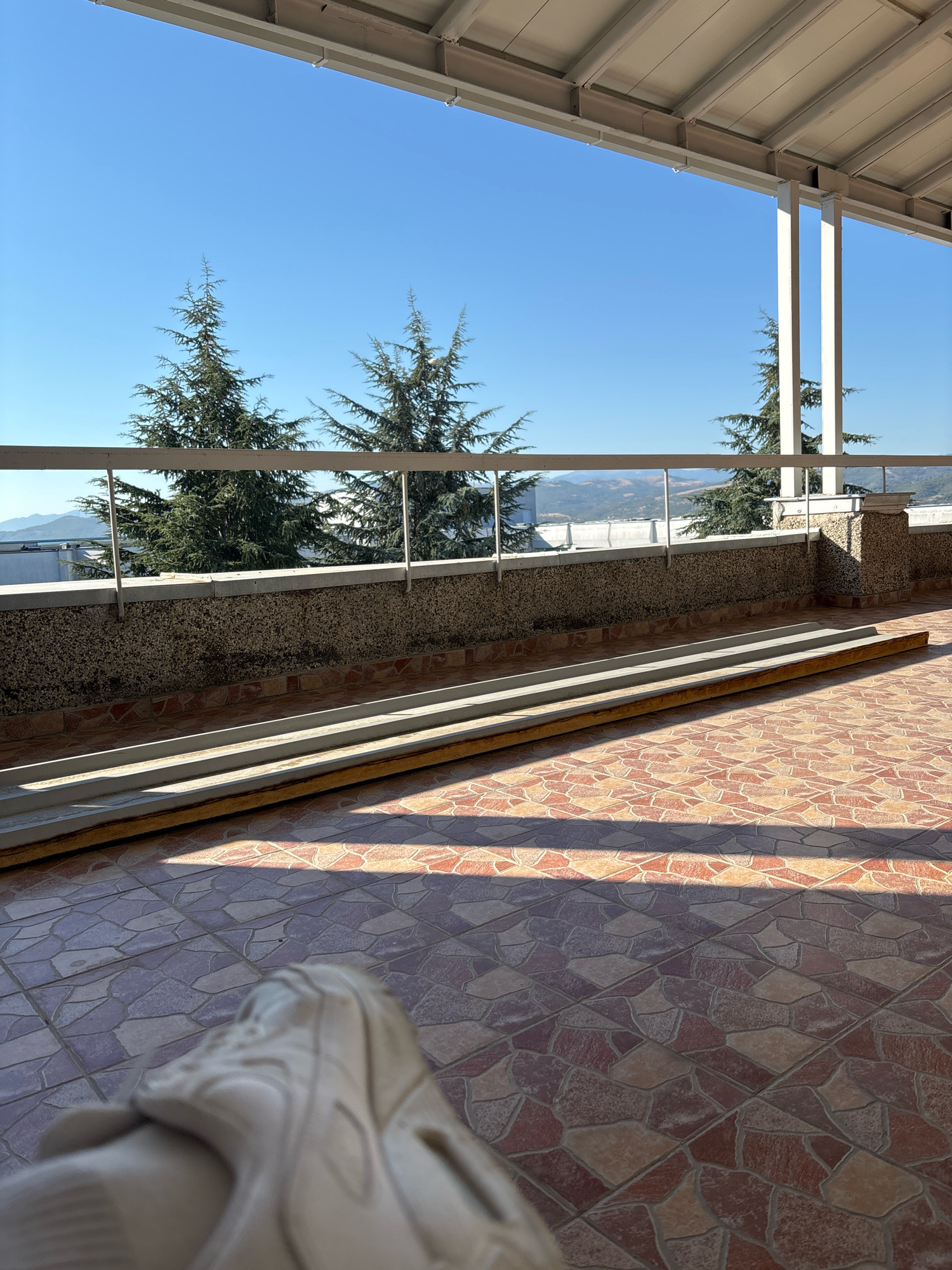 The image shows a view from a covered balcony or terrace. In the foreground, partially visible, is a person's foot wearing a light-colored athletic shoe. The floor of the balcony is tiled with irregularly shaped terracotta-colored tiles. Along the edge of the balcony is a railing made of metal poles and a horizontal bar. Beyond the railing, there is a wall constructed of rough, speckled material, topped with a concrete coping. Behind this wall, several tall evergreen trees are visible. In the background, a clear blue sky dominates, with a distant landscape of rolling hills and some scattered buildings. The overall impression is of an outdoor space in a warm, sunny climate. This scene is likely located in the city of Blagoevgrad, Bulgaria, given its typical terrain and vegetation.
