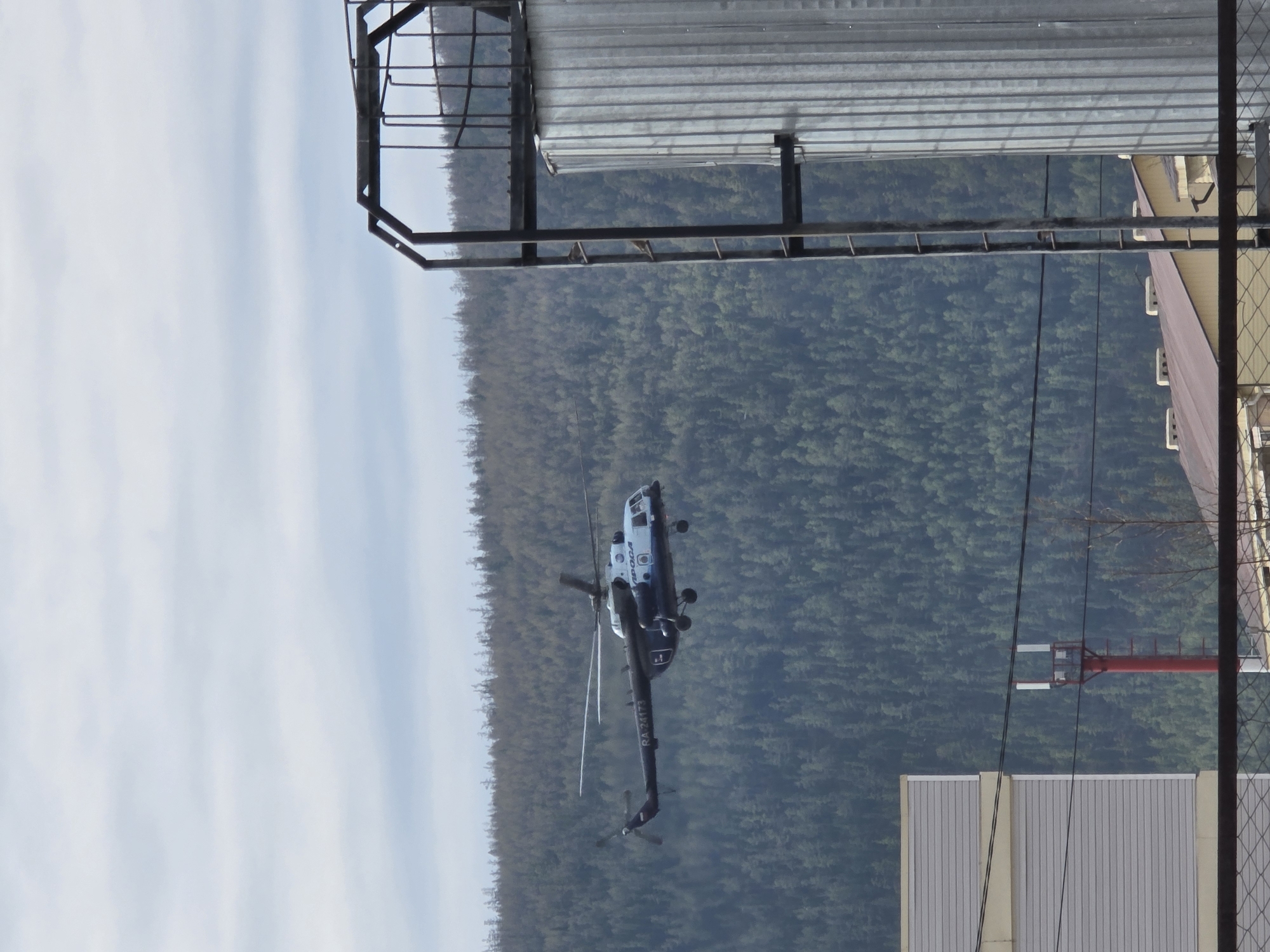 A helicopter is airborne near Lensk, Russia, with a dense forest visible in the background. Utility poles and building structures are in the foreground. The sky appears partly cloudy. The helicopter bears markings "APOGA" and "RA-24173".