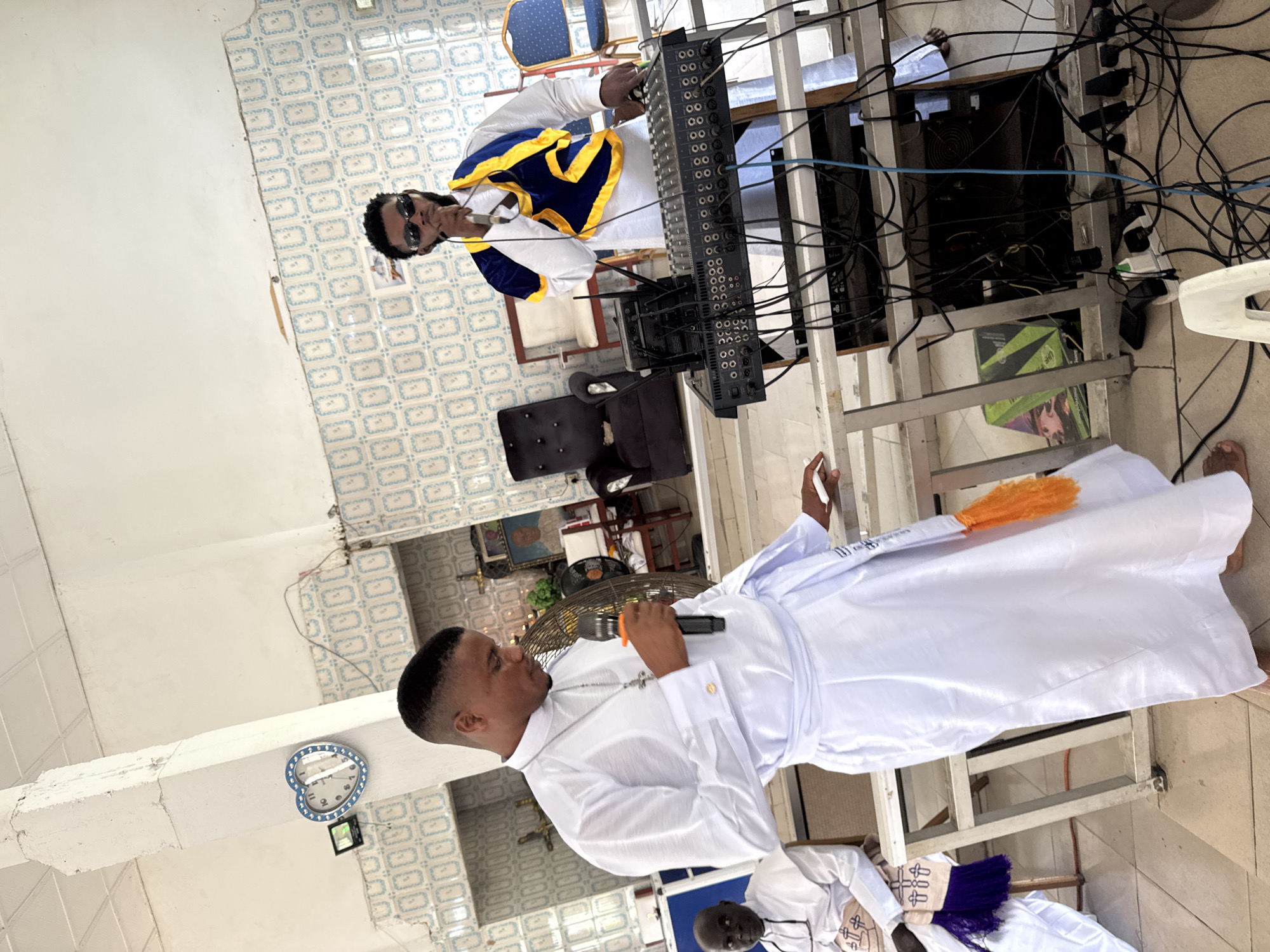 The image depicts a scene inside a building, likely a place of worship, with a distinctive tiled wall featuring a repeating pattern of blue and white squares and circles. In the foreground, two men are prominently featured. The man on the left, dressed in a flowing white robe with a silver cross necklace, holds a microphone and appears to be speaking. He has a focused expression and his gaze is directed downwards. He is standing beside a metal railing.
To the right, another man, wearing a white robe with a blue and gold sash, holds a microphone to his lips and is singing or speaking into it. He is positioned behind a sound mixing board, which is a complex arrangement of black electronic equipment with numerous knobs, buttons, and cables. This man is wearing dark sunglasses and has a beard.
In the background, other individuals can be seen, including a man seated in a white robe on the left, and various chairs and furniture, including a large black armchair. A heart-shaped clock with a blue border is visible on the wall near a white pillar. The overall lighting suggests an indoor environment with some natural light, as indicated by the shadows. The tiled floor is visible in the foreground. The environment appears to be a public space, possibly a church or a community hall.