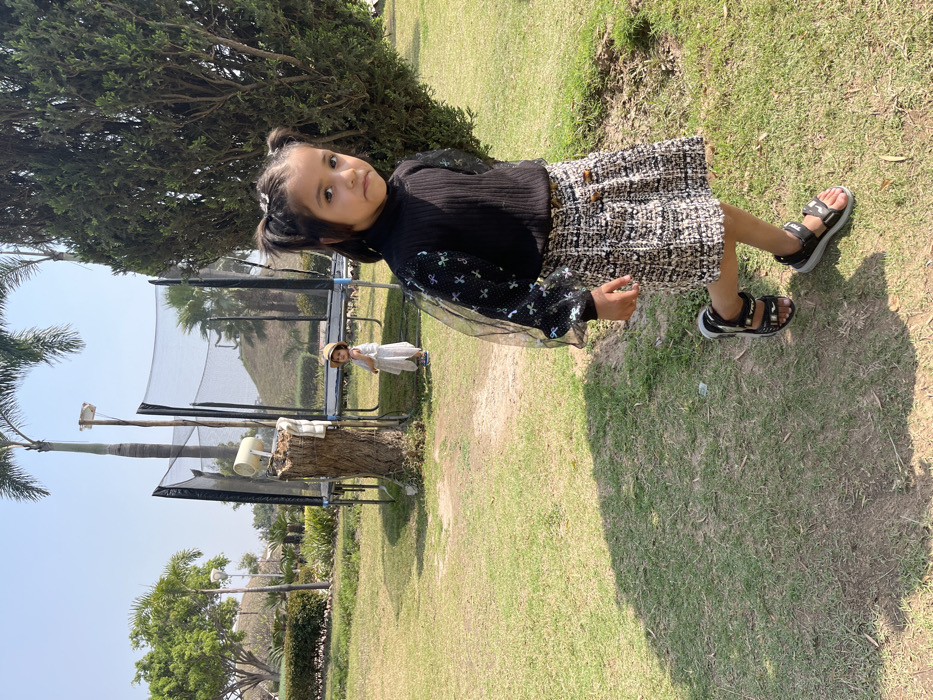 The image depicts two young girls in a park in Sahibzada Ajit Singh Nagar, India. The girls are both wearing casual clothing. The girl in the foreground is wearing a black sweater with a white and black checkered skirt. She has her hair pulled back in a ponytail. The girl in the background is wearing a white dress and a straw hat. She is standing near a trampoline. Both girls are walking on a grassy lawn. The scene is daytime and the weather appears sunny. There is a small tree in the background. The trampoline is in the middle of the background, with a large tree to the left. The girls appear to be in a playful mood.