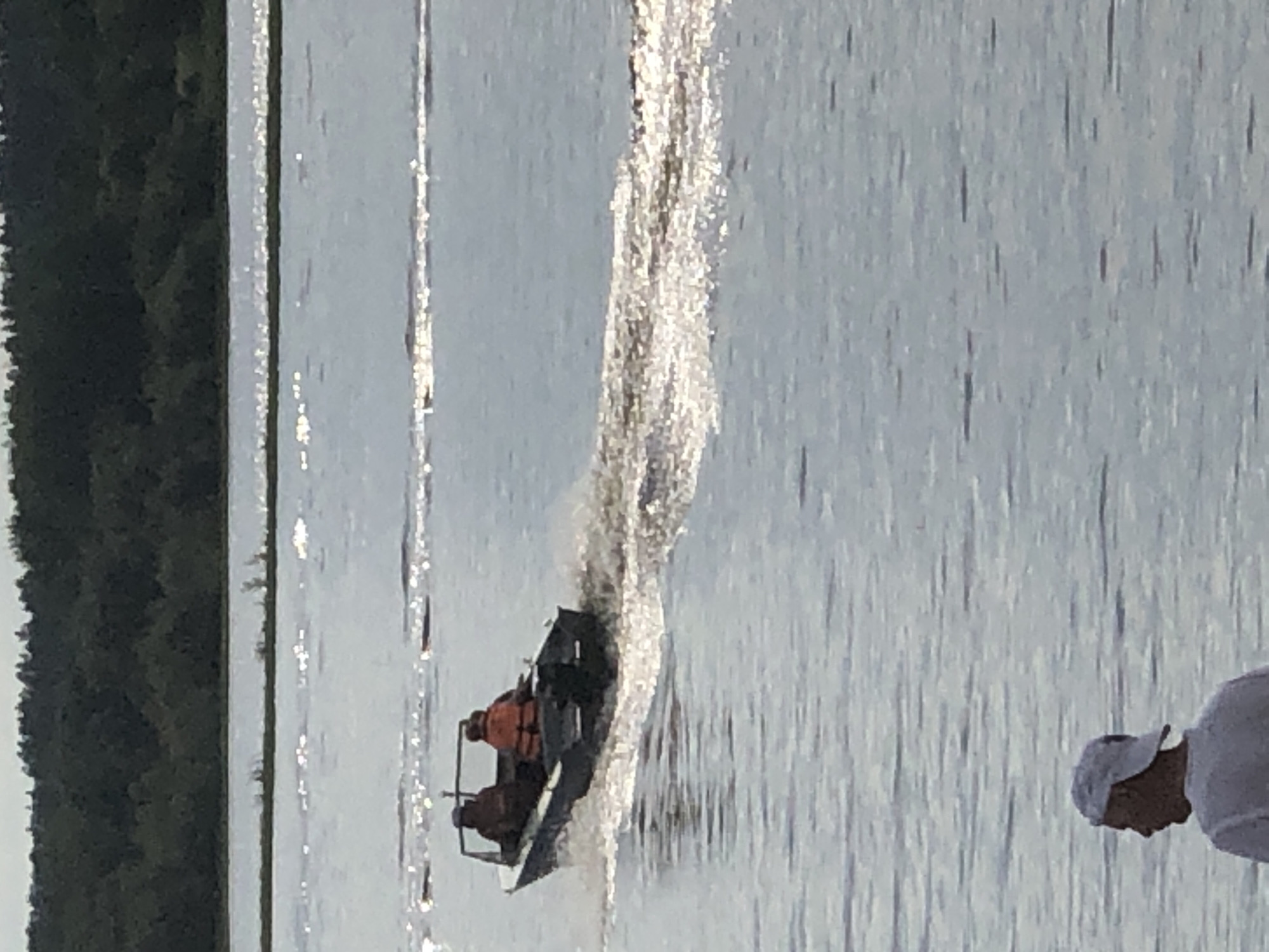 A small, dark-colored motorboat with two occupants is moving on a body of water. The person in the front is wearing a dark hat and a dark shirt, while the person in the back is wearing an orange life jacket. The boat is creating a wake behind it. In the foreground, a person wearing a white baseball cap and a white shirt is standing and looking away from the camera, facing the water. In the background, a dense line of dark green trees borders the water. The sky is overcast. The scene appears to be a recreational area, possibly a lake or river, in Solikamsk, Russia.