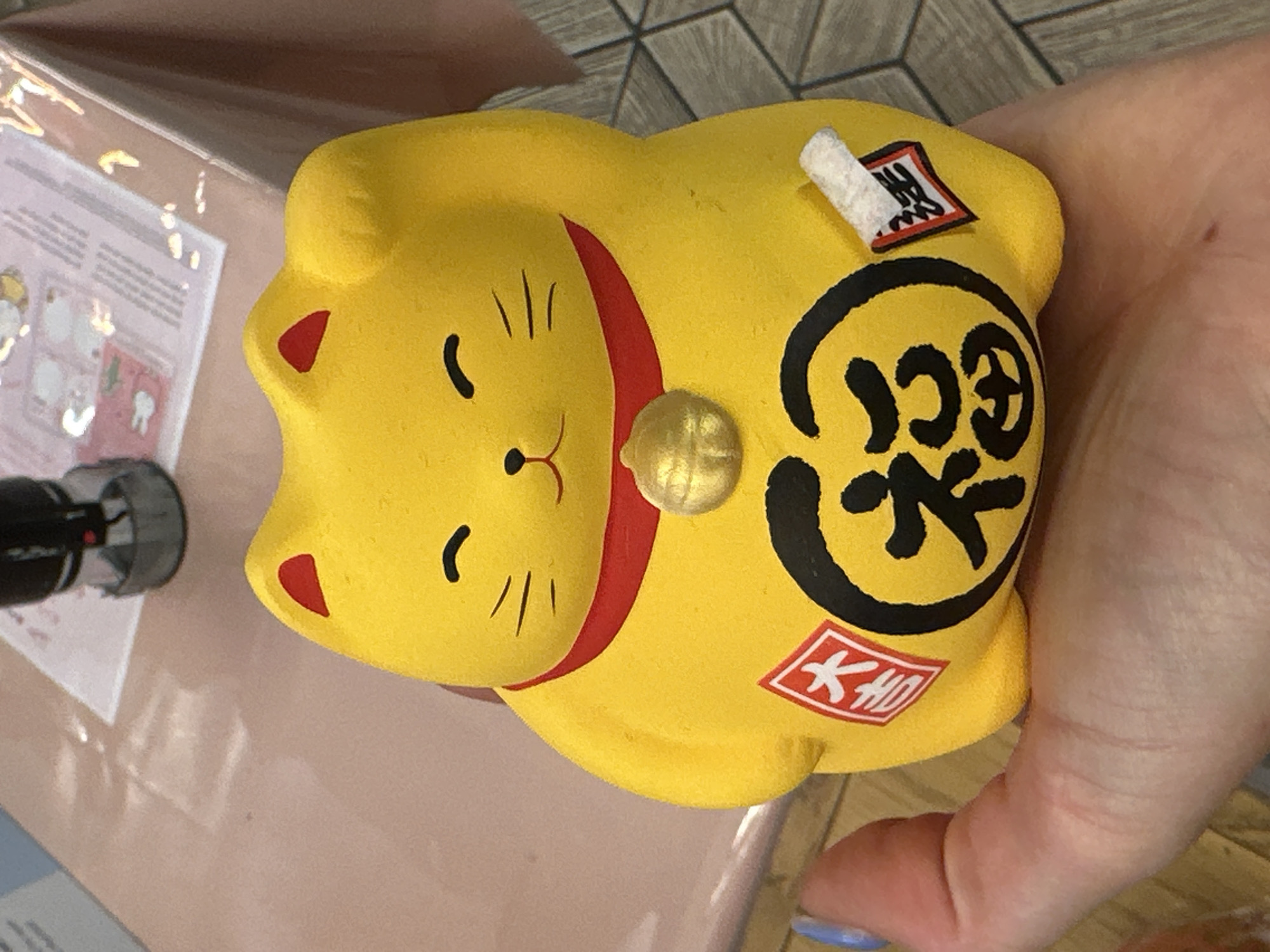 A yellow Maneki Neko figurine is held in a person's hand. The figurine has a red collar, gold bell, and Japanese characters on its body. The surrounding environment appears to be an indoor setting with a brochure or package visible on a nearby surface. No specific location in London is evident.