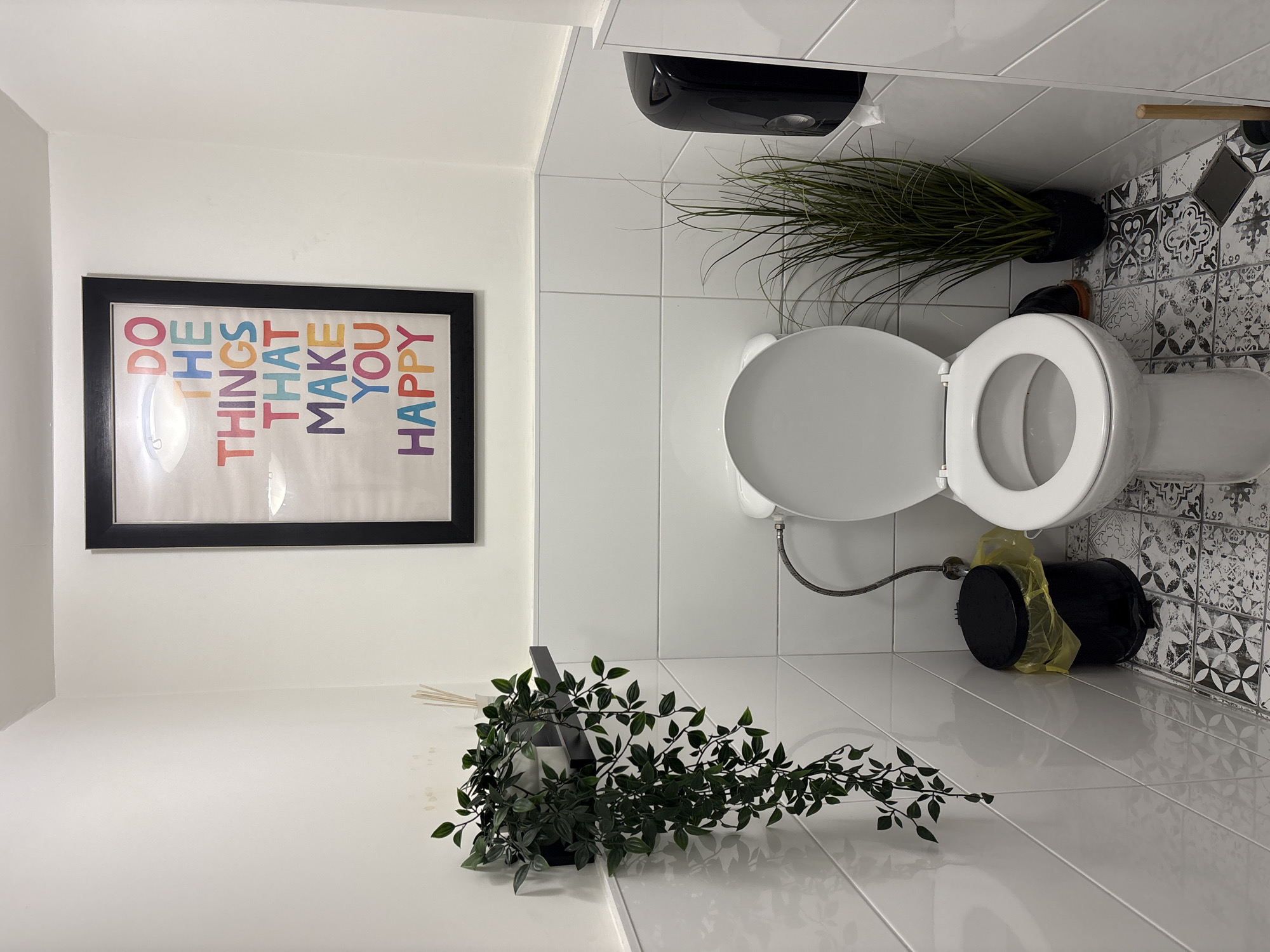 A bathroom interior is depicted. A white toilet with an open lid sits on patterned tile flooring. A black trash bin with a yellow liner is near the toilet. Above the toilet is a wall-mounted black dispenser and a potted green plant. Adjacent to the toilet, a framed poster reads "DO THE THINGS THAT MAKE YOU HAPPY." A green plant is on a white surface. The location is Veliko Tarnovo, Bulgaria.