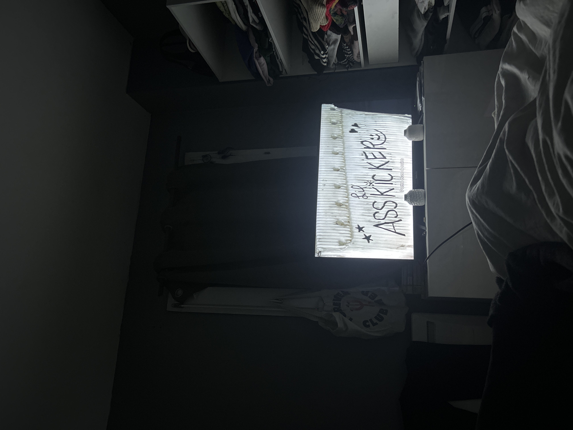 The image shows a dimly lit room. A white closet is visible at the top of the image, with clothing hanging inside. Below the closet, a television screen displays the word "ASSKICKER!" with decorative elements. Next to the television screen is a white bedside table, with rumpled white sheets. A white reusable bag with the word "CLUB" can be seen. The setting appears to be a bedroom. The image provides no information about Lannoy, France.