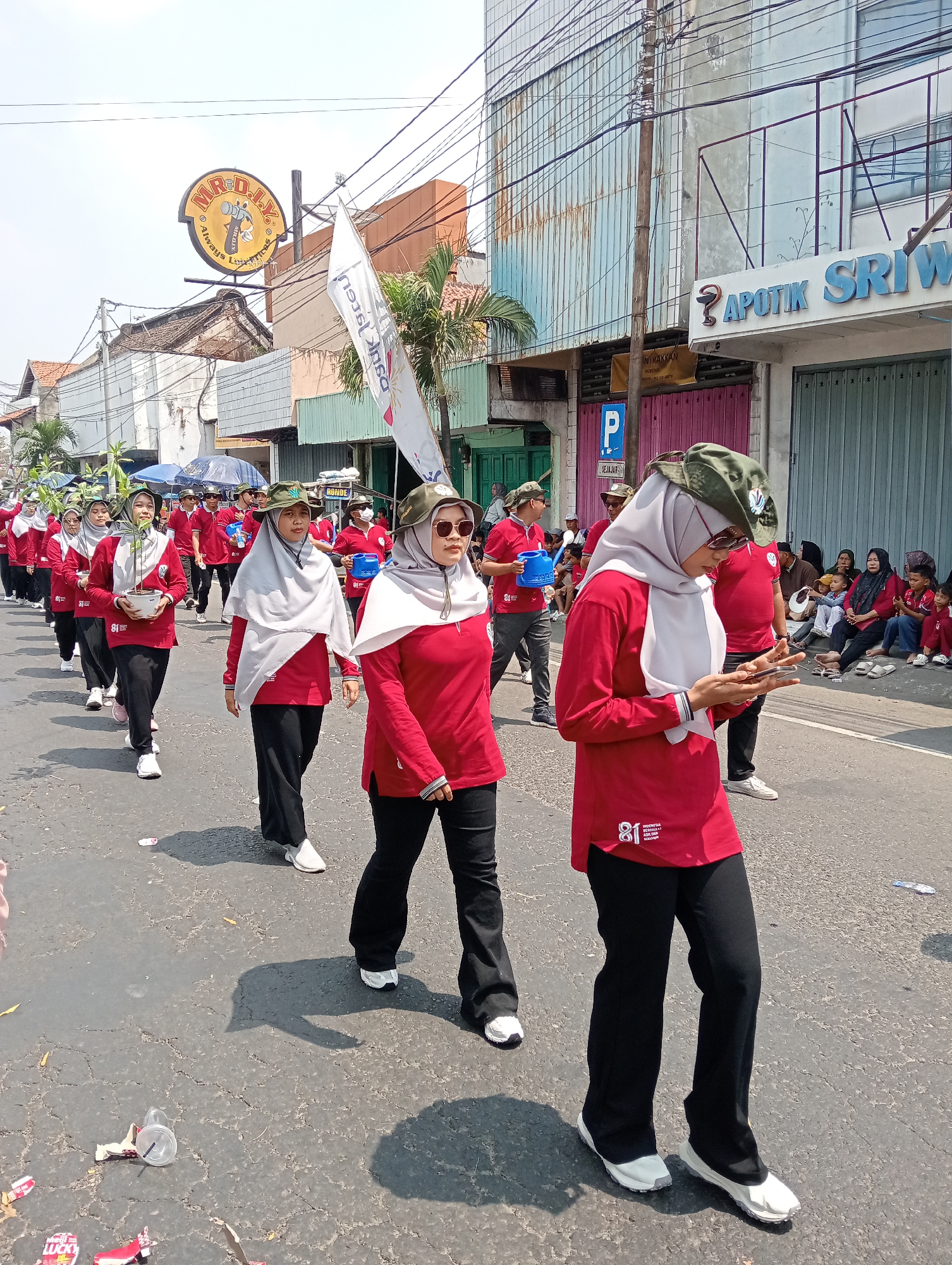 A group of people, likely participating in a parade or procession, walk down a street in Demak, Indonesia. The individuals are mostly wearing matching red shirts and black pants, with some wearing headscarves and hats. Several people are holding small potted plants. In the background, there are buildings, including a shop with a prominent sign for "APOTIK SRIWI" and another sign with a logo featuring a hammer. The street appears to be paved with asphalt and shows signs of wear. The sky is bright and clear, suggesting a sunny day. There are some spectators visible on the side of the street. Trash, including plastic cups and wrappers, is scattered on the ground.