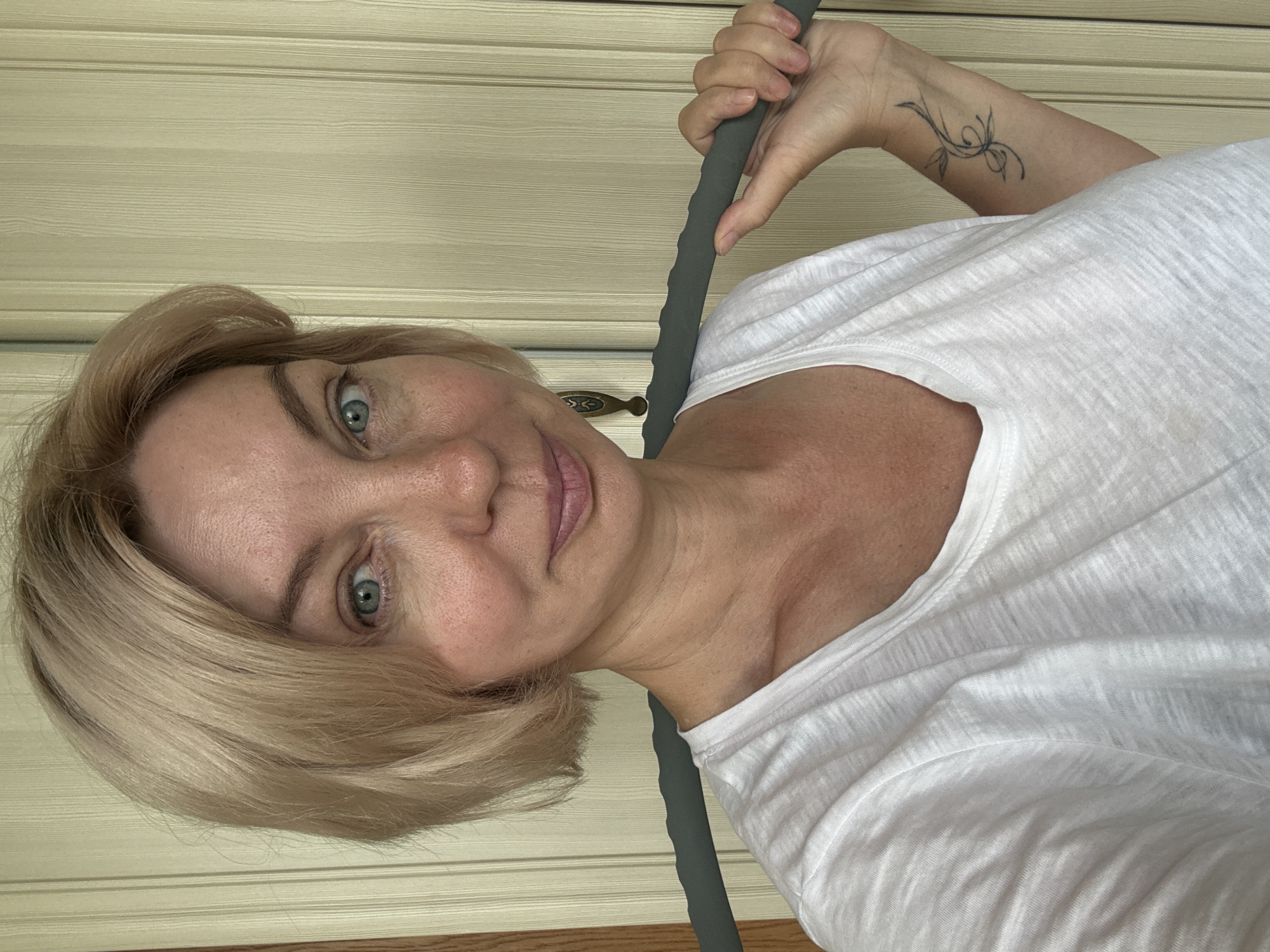 A middle-aged woman with blonde hair is captured in a close-up, shoulder-up selfie. She is wearing a white t-shirt and holding a grey, ribbed massage roller across her shoulders. The woman has light blue eyes, fair skin with freckles, and is smiling slightly. A small tattoo of a swirling vine is visible on her left forearm. The background consists of light beige vertical blinds and a sliver of a wooden door frame. The lighting suggests an indoor setting, possibly a home in Sochi, Russia.