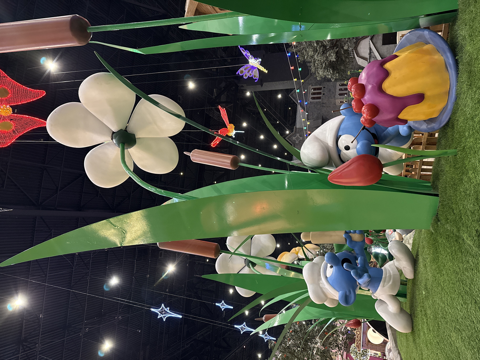 The image depicts a Smurf-themed display, possibly a holiday or promotional installation, located in Moscow, Russia. The display features oversized cartoon figures of Smurfs, one with a chef's hat holding a dessert topped with red fruits, and another in a similar outfit hanging upside down from what looks like a giant plant. The scene is set indoors, with a dark ceiling decorated with star-shaped and dragonfly-shaped illuminated ornaments. The ground is covered in artificial turf. In the background, building facades and festoon lights are visible. Overall, the image captures a whimsical and playful scene, likely aimed at attracting visitors and creating a festive atmosphere.