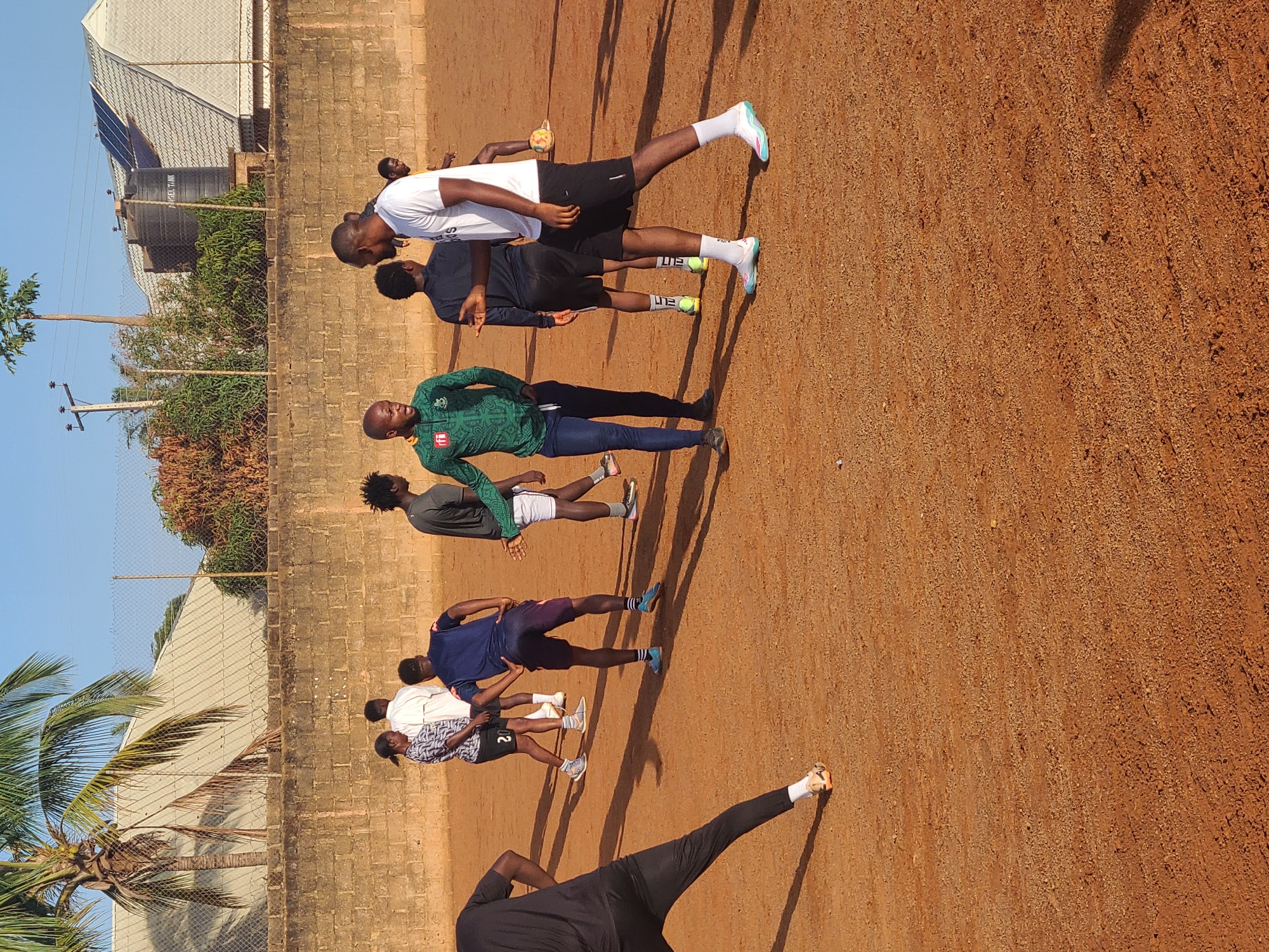 This image, captured outdoors and rotated 90 degrees clockwise, displays a group of approximately eight to nine young men standing on a reddish-brown dirt or sand surface. The men, dressed in various athletic attire including t-shirts, shorts, and long pants, appear to be engaged in a sports training session or warm-up. Some are standing in a line, while others are slightly scattered, with one individual in the foreground (left after correction) posing with hands on hips.
Notable details include a small, light-colored ball near one person's foot. Many of the men wear athletic shoes and socks, with the word "ELITE" visible on some of the socks. One individual's black shorts display the number "20." In the background, a low brick wall and a chain-link fence line the perimeter of the field. Beyond this, several buildings with light-colored roofs are visible, one featuring multiple solar panels and a large black water tank. Partial text "RAGGEI TANK" is discernible on the water tank. Lush green trees, including a prominent palm tree, frame the scene against a clear blue sky. The strong, distinct shadows cast by the individuals indicate bright, direct sunlight, suggesting it is either morning or late afternoon. The overall setting is an open, sunny athletic ground in a warm climate.
