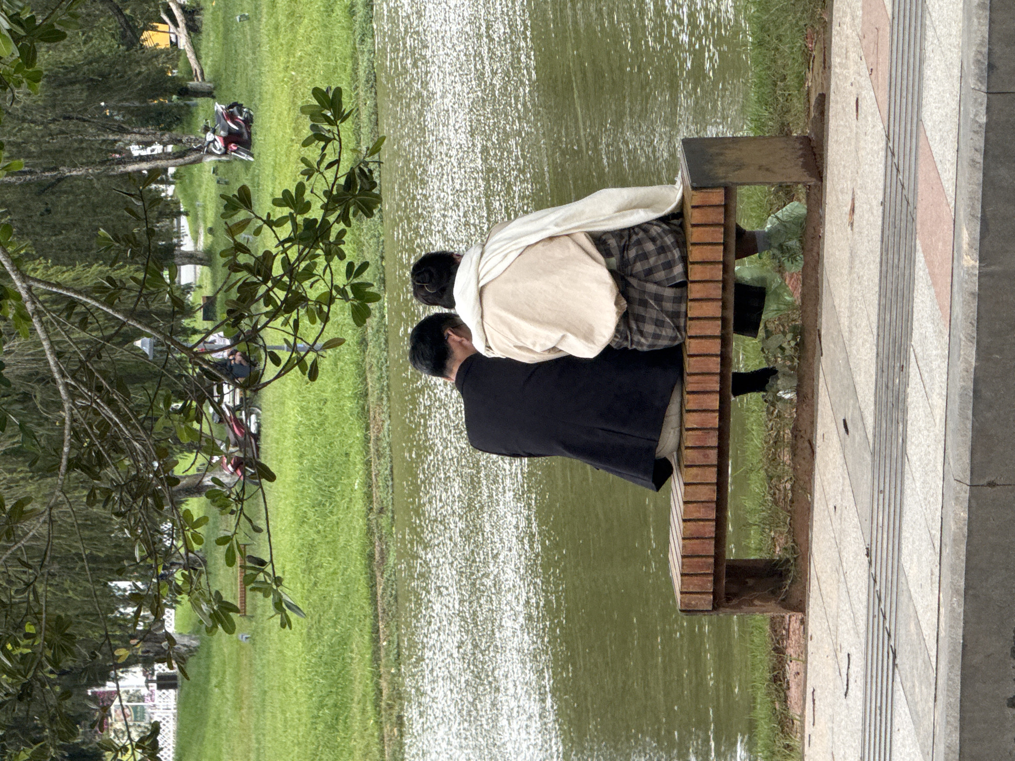 In Da Lat, Vietnam, two individuals, viewed from behind, are seated on a wooden bench by a body of water. The person on the left wears a black jacket and light-colored pants. The person on the right is draped in a light-colored shawl over a tan top, and patterned plaid pants. Both individuals are wearing light green shoe covers. The bench is made of wooden planks and has dark metal legs, situated on a paved walkway. Beyond the walkway and bench, a grassy area slopes down to the water, with trees providing shade. In the background, more trees are visible, and a red scooter is parked on the grass. The water's surface is rippled, reflecting light.