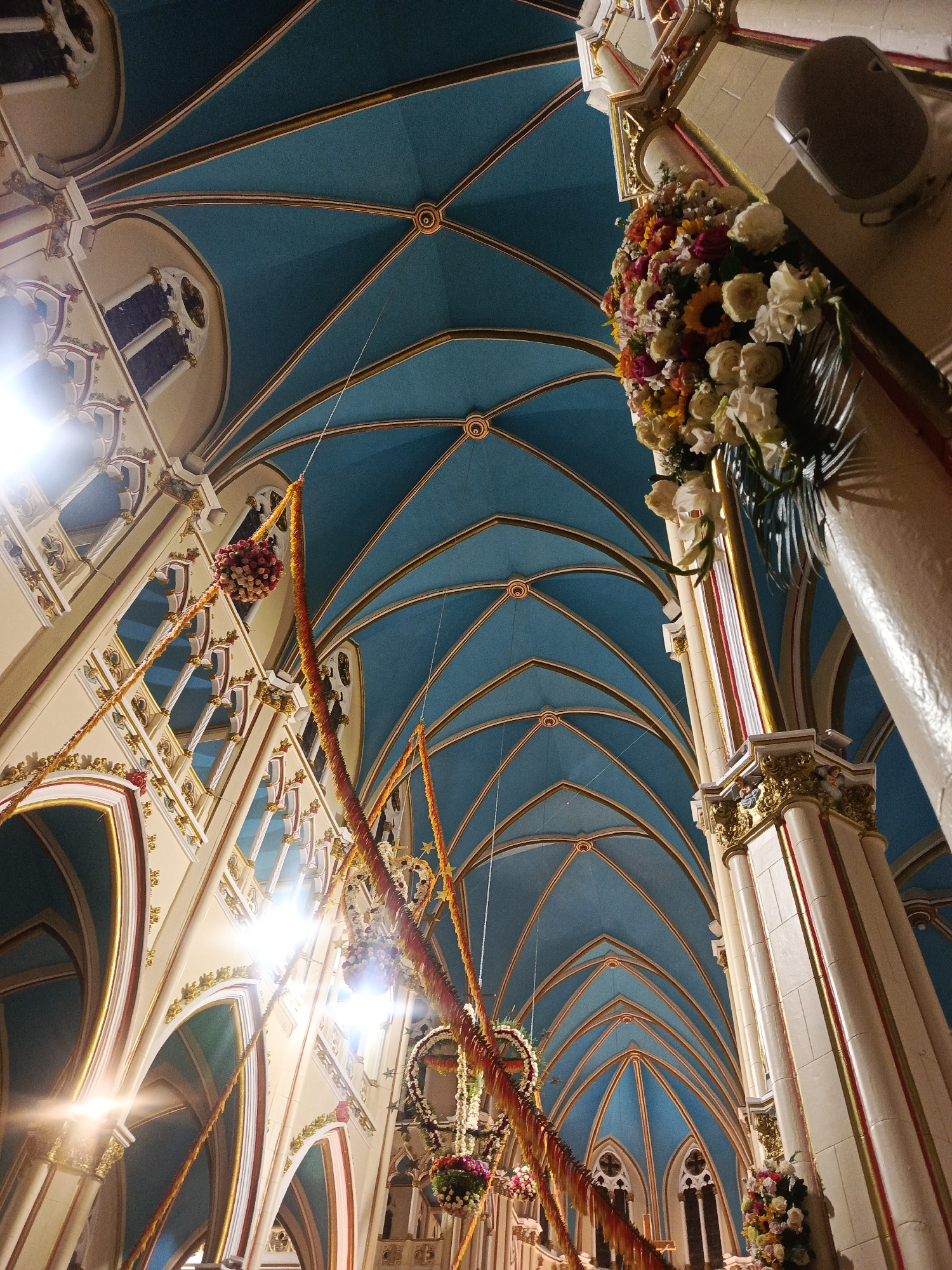 The image shows the interior of a large church in Unknown, Unknown. The ceiling is a series of arches painted a deep blue, accented with gold trim. Pillars supporting the structure are decorated with white and gold paint, and ornate carvings. A large floral arrangement adorns one of the pillars. Festive decorations, consisting of orange and white garlands, hang from the ceiling and arches. Windows line the walls, allowing natural light to filter into the space.