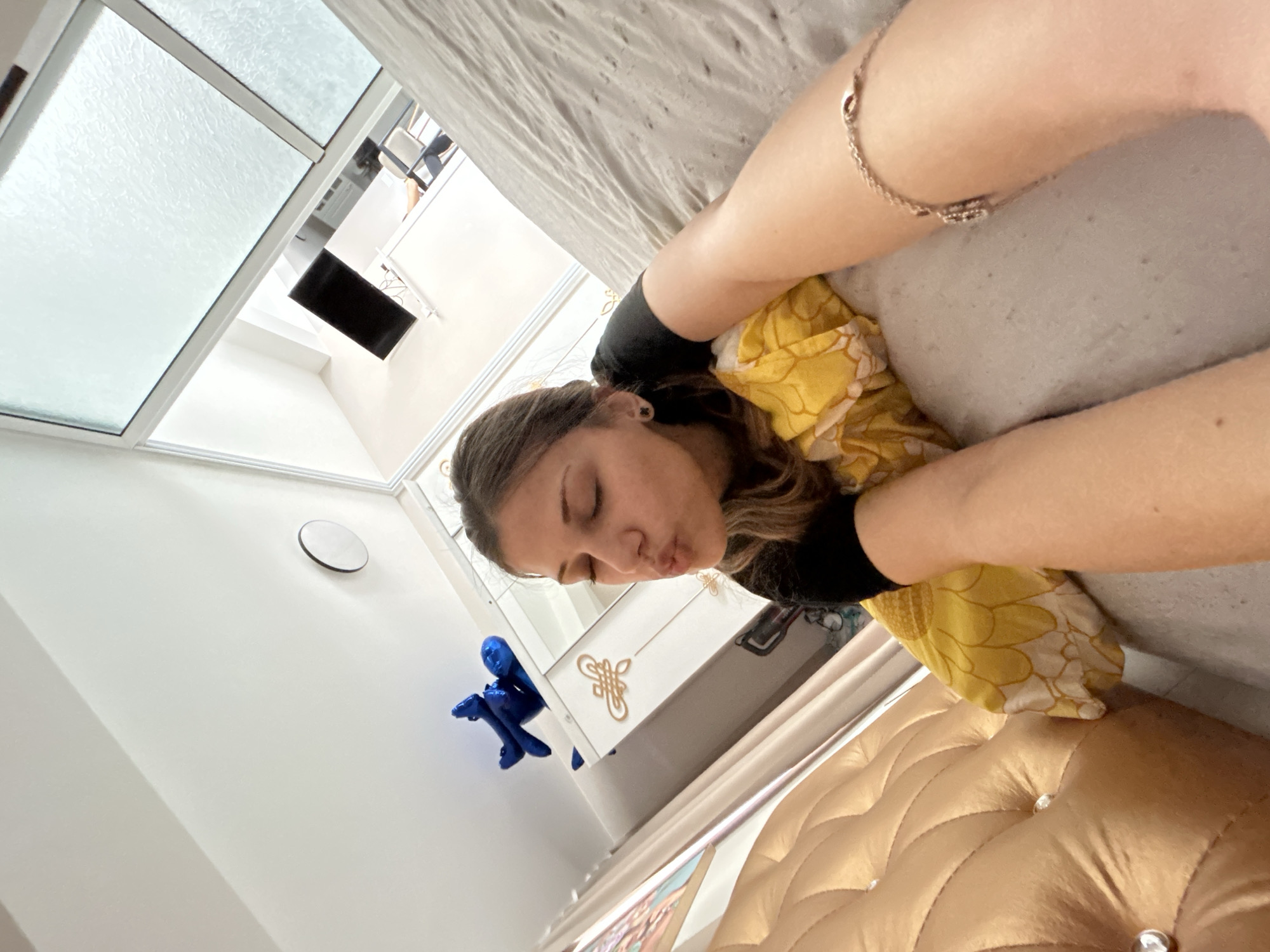 A female subject is captured in a selfie, lying on her side on a bed within an interior room. She has brown hair, wears a black short-sleeved top, small black clover-shaped earrings, and a silver chain bracelet on her right wrist. Her eyes are closed, and her lips are pursed in a kissing gesture, with her head resting on a yellow pillow featuring a white floral pattern. The bed includes a gold-colored, tufted headboard adorned with clear crystal buttons and is covered by a greyish sheet or duvet. In the background, a white wardrobe with gold ornamental handles is visible. A blue foil balloon, shaped like a stylized human figure, is positioned near the ceiling. A circular white ventilation cover is mounted on a white wall. Above, a skylight window with textured glass reveals a glimpse of another room or floor, showing a white staircase railing, a dark rectangular object (possibly a screen), and part of a chair. The scene is an interior room, likely a bedroom, within a residential dwelling in Sofia, Bulgaria.