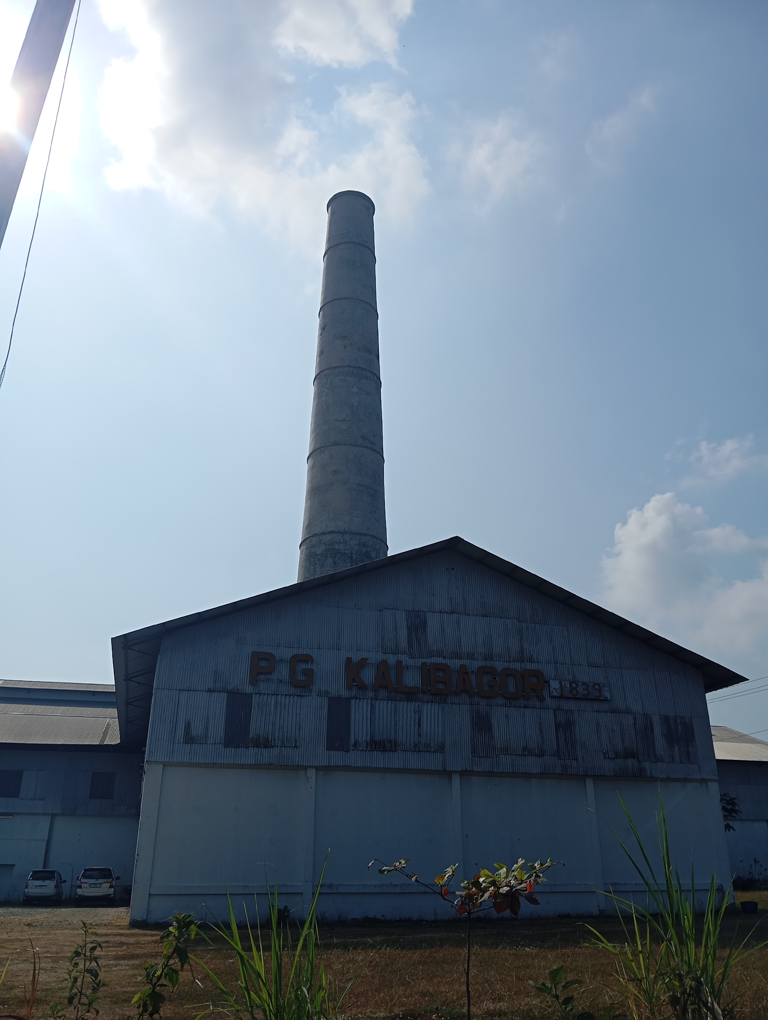 The image captures a daytime view of a large, industrial building in Sokaraja, Indonesia. A tall, cylindrical smokestack, constructed of segmented concrete rings, dominates the skyline, positioned just behind and to the left of the building's gable roof. The building itself is clad in corrugated metal siding and features a white lower facade. Large, dark letters spelling "P.G. KALIBAGOR" are visible across the front of the building, with the number "1839" following. To the left of the main building, a portion of another structure with a similar roofline and corrugated metal siding is visible. Two cars are parked on a paved area in front of the structures. The foreground is characterized by dry grass and some sparse green vegetation, including a small sapling with reddish-orange leaves. The sky above is a clear blue, dotted with a few white clouds, and the sun is bright, casting a lens flare on the left side of the image.
