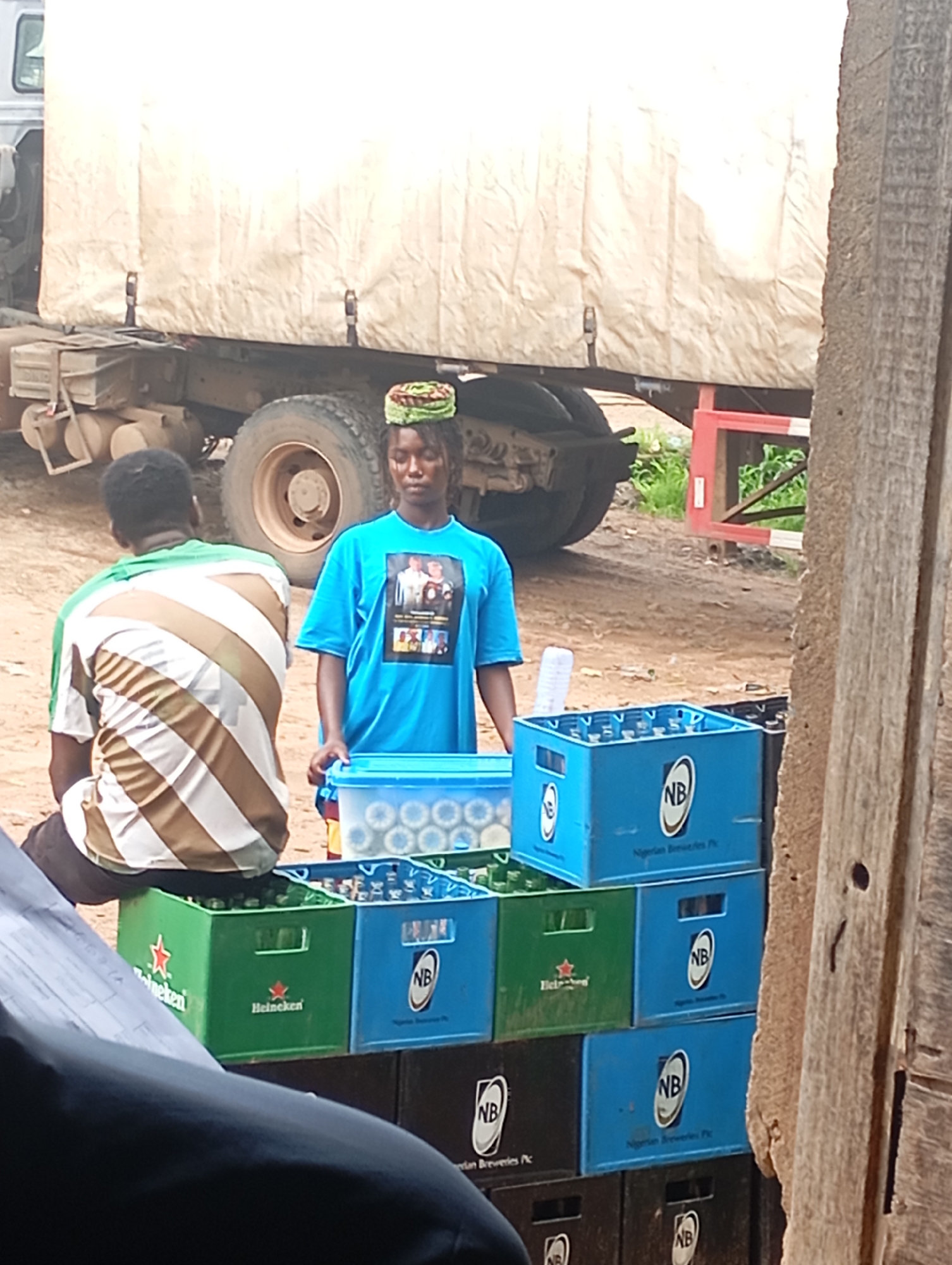 In Unknown, Unknown, a woman wearing a blue t-shirt and a green headwrap stands beside stacks of bottled beverages. The t-shirt displays a framed image. She is looking down. To her left, a person wearing a brown and white striped shirt is seated with their back to the camera. In front of them are crates of drinks, including green Heineken crates and blue "NB Nigerian Breweries Plc" crates. Behind the people and crates is the rear section of a large, tan-covered truck with its tarp pulled taut and securing straps visible. The ground appears to be dirt and gravel, with some sparse green vegetation. The scene is outdoors, with a wooden structure visible on the right side of the frame.