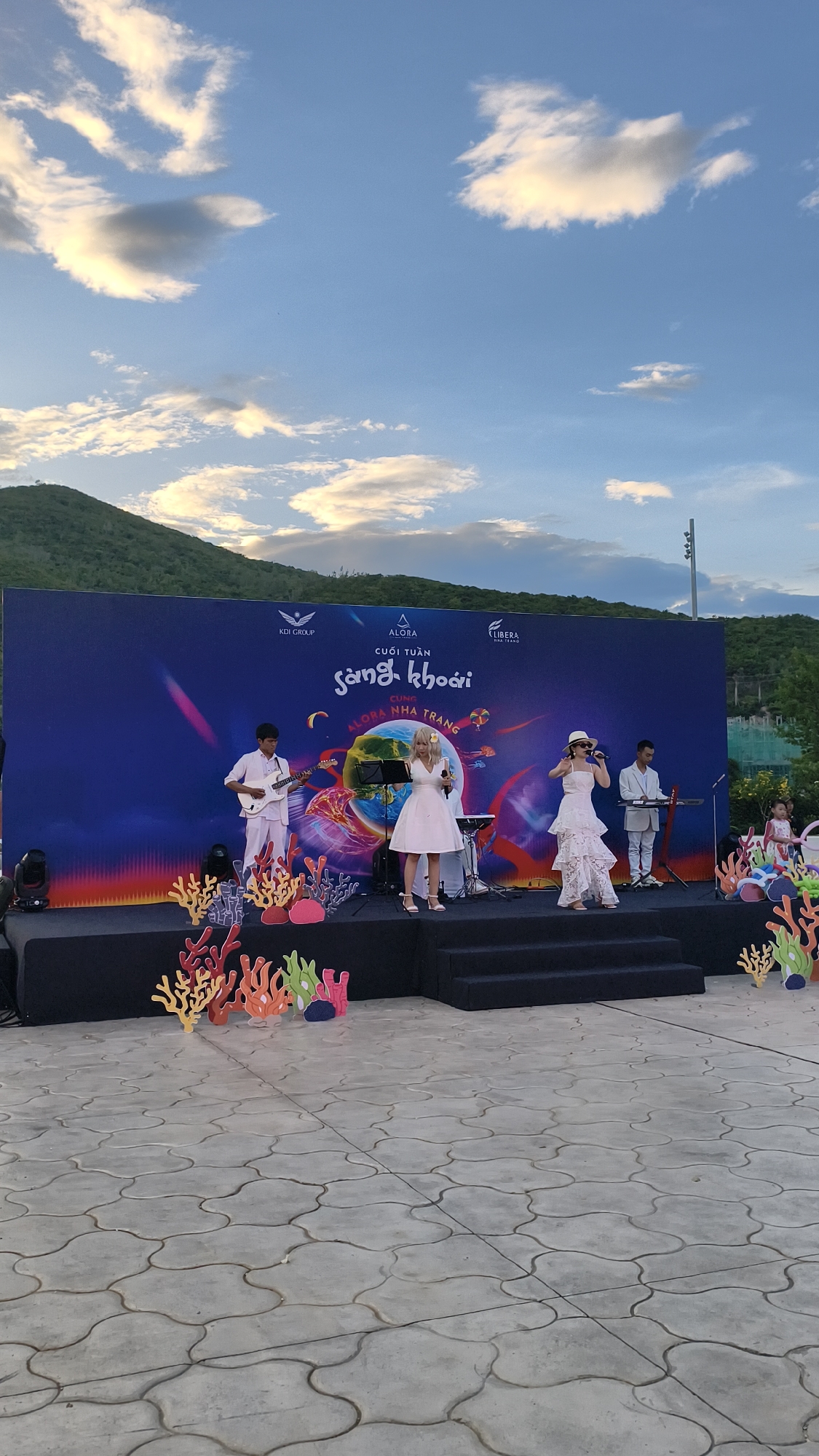 The image depicts an outdoor stage event set against a backdrop of a large banner and a scenic hillside. The banner displays Vietnamese text that translates to "Weekend Fun with Alora Nha Trang," featuring logos for KDI Group, Alora, and Libera. The stage itself is adorned with colorful, cartoonish coral decorations. On stage, four musicians are performing. A young man in a white outfit plays a white electric guitar, and two female singers are in the center. One singer, wearing a white tiered dress and a hat, is singing into a microphone, while another singer in a white dress with a microphone stands beside her. A third musician, in a light blue suit, plays a keyboard on the right side of the stage. In the background, a natural hillside covered in lush greenery rises under a cloudy sky with hues of blue and white. The ground in front of the stage is paved with large, interlocking grey tiles. To the right of the stage, a few spectators are visible, including children. The overall scene suggests a festive evening event likely held in a coastal area of Nha Trang, Vietnam, possibly an entertainment venue or public space associated with the Alora brand.