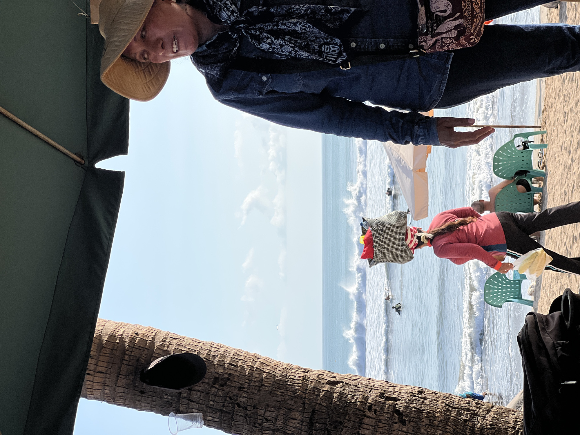A woman wearing a wide-brimmed hat and a denim shirt stands on a beach in Kuta, Indonesia, looking to her right. A patterned scarf is tied around her neck. A purse with an elephant design is slung across her body. To her left, a portion of a palm tree trunk is visible, with a dark object hanging from it. In the background, the ocean has waves, and a few people are surfing. Another woman, wearing a red shirt and black pants, walks towards the viewer, carrying a bag of groceries. A green plastic chair and a beach umbrella are also in the background. The sky is clear and blue.