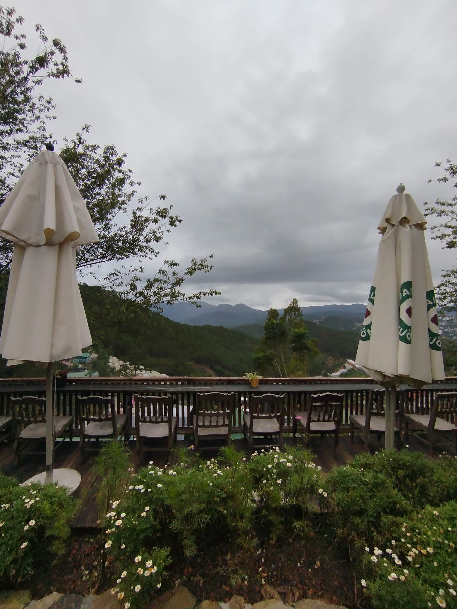 The image shows a view from a balcony in Xuan Truong - Da Lat, Vietnam, with wooden chairs lined up along a railing. Beyond the railing, there is a landscape of rolling hills and mountains under an overcast sky. Two closed white umbrellas stand on either side of the chairs. Below the railing, there are bushes and flowers in the foreground.