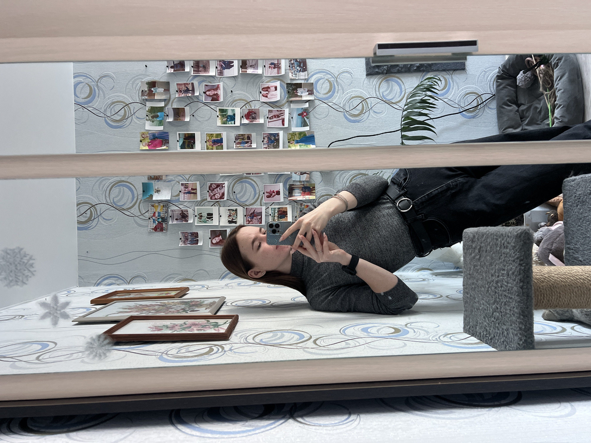A person lies on a surface that appears to be a bed with three framed objects on it, taking a photo with a smartphone. The bed's surface is patterned with white and blue swirls. The person is wearing a grey long-sleeved shirt and dark pants with a belt. To the right, partially visible, is a grey cat tree. Behind the person, attached to the wall, are numerous small photographs arranged in rows. The wall has a patterned wallpaper with blue and grey swirls. A plant and a grey pet bed are situated further back along the wall. The photograph is taken as a reflection in a mirror, and the mirror frame is visible. No information about the city of Chaikovsky, Russia is detectable in the image.