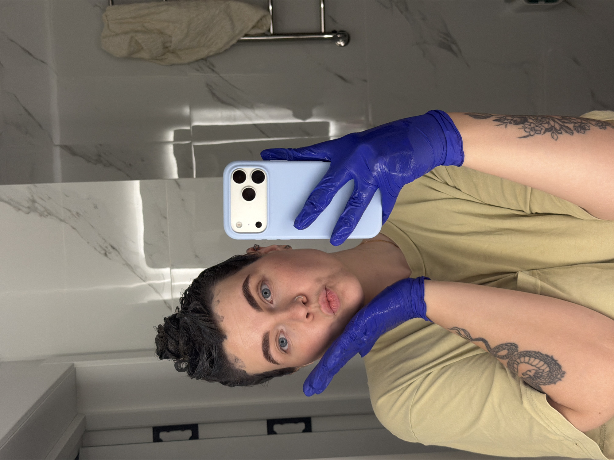 A person with blue eyes and dark hair is taking a selfie in a bathroom. They are wearing blue gloves and a tan shirt. Their hair appears to be freshly dyed with black hair dye. The person has a tattoo on their arm. The bathroom has white tiled walls and a towel rack. There is no indication of the city Chaikovsky, Russia.