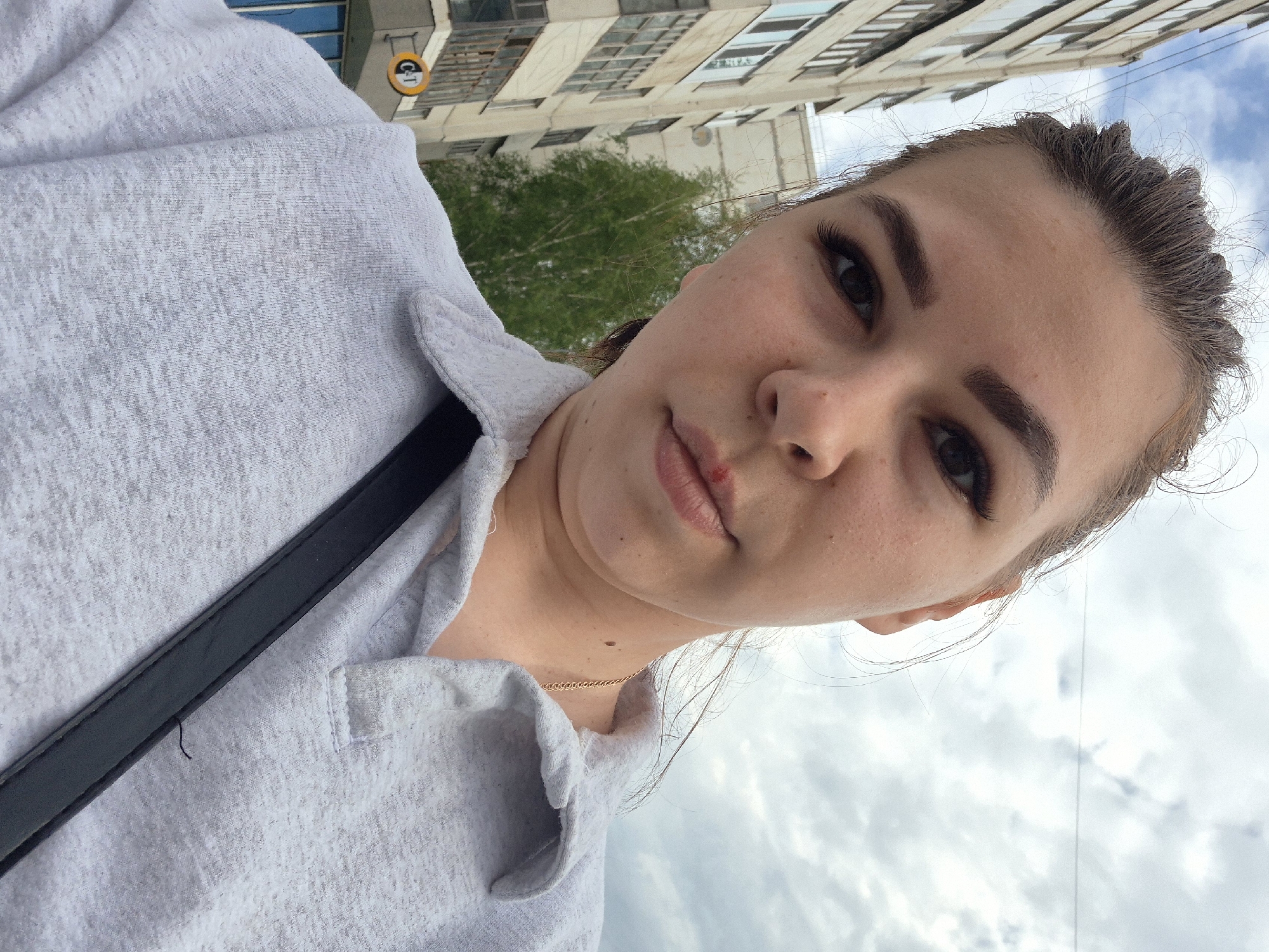 A close-up, selfie-style photograph depicts a fair-skinned young woman with brown hair pulled back, wearing a light gray collared shirt and a black shoulder strap. A minor abrasion is visible on her lower lip. Behind her, out of focus, is an urban scene of buildings and a green tree against a cloudy sky. A yellow and black sign is partially visible on the building, indicating the location as Tobolsk, Russia.