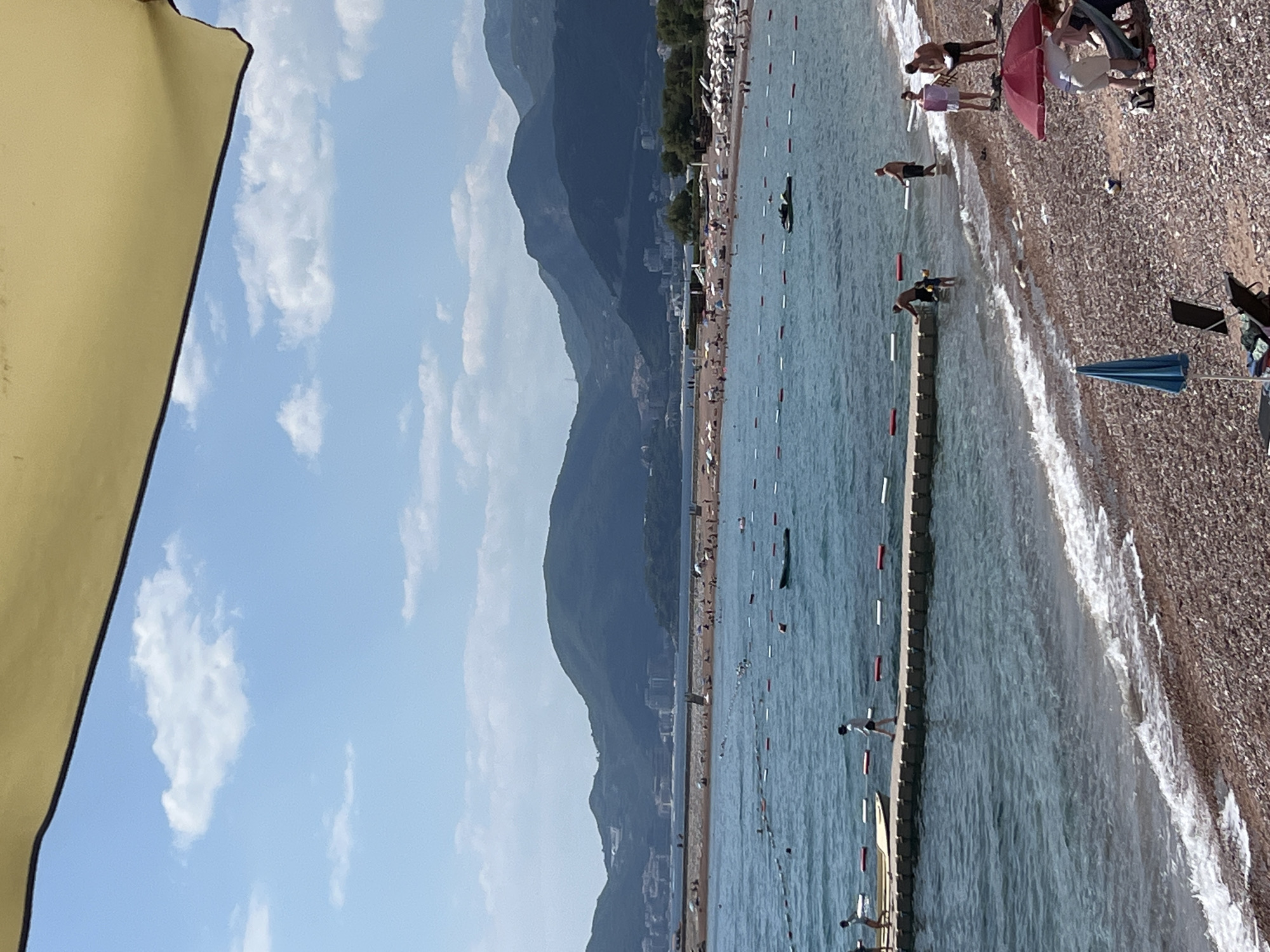 This is a photograph taken at a beach in Sveti Stefan, Montenegro. The image is oriented vertically. In the foreground, a large portion of a yellow umbrella or awning is visible on the left, creating a partial frame for the scene. The background reveals a crowded beach with many people relaxing and swimming in the clear blue water. The beach itself is composed of small pebbles.
The water is calm with gentle waves lapping at the shore. A white barrier with red markers separates the swimming area from the deeper water, and a long, low wooden structure extends into the sea, possibly a pier or a floating platform. Several individuals are visible in the water, some swimming and others wading near the shore. On the beach, people are scattered, some sitting on towels or chairs, while others are standing or walking.
In the distance, a line of buildings and trees forms the backdrop to the beach, suggesting a developed coastal area. Towering behind the coastline are majestic, dark green mountains, their slopes partially covered in shadow, indicating the sun is likely shining from behind them or to the side. The sky is a bright, clear blue with a few scattered white clouds.
The overall atmosphere appears to be one of leisure and a pleasant summer day. The presence of numerous people suggests it is a popular tourist destination, likely during peak season. The sunlight appears to be relatively bright, casting some shadows, which could place the time of day as mid-morning or afternoon. There is no visible text in the image.