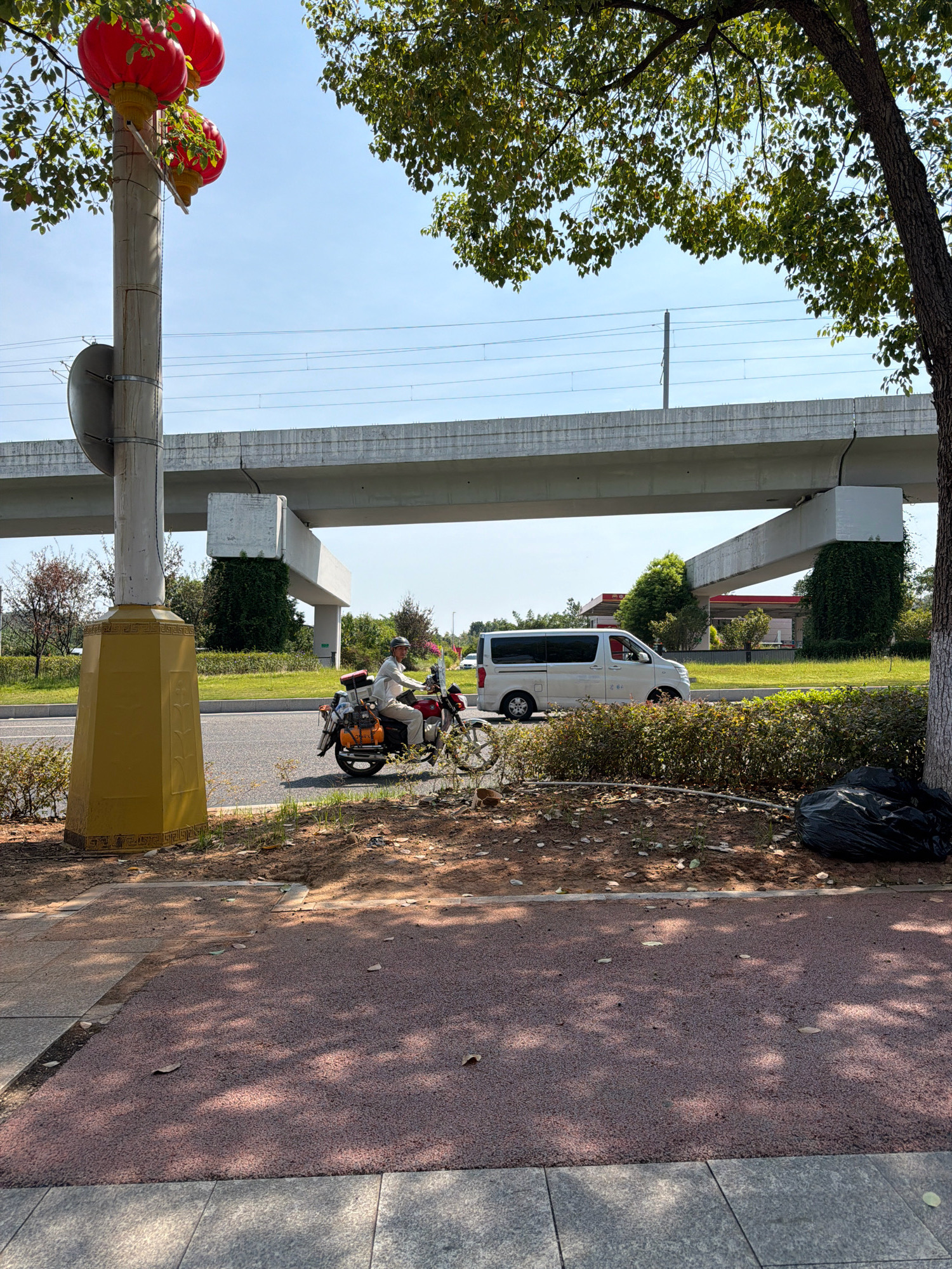 In Jiangjin, China, on a clear day, a motorcycle rider wearing a helmet and a light-colored shirt navigates a road beneath an elevated structure. The motorcycle is red and adorned with luggage, including an orange bag and a red case. To the right of the motorcycle, a white van is visible, also on the road. The road is flanked by a grassy area and a row of green bushes. A light pole with red lanterns hangs on the left side of the image, partially obscuring a grey, oval-shaped object. The elevated structure above appears to be a bridge or overpass, with concrete pillars supporting it. A dark trash bag lies on the ground near the bushes on the right. The foreground shows a red, textured pathway and a grey tiled sidewalk, with shadows of tree leaves dappled across them. Lush green trees and foliage are present throughout the scene.