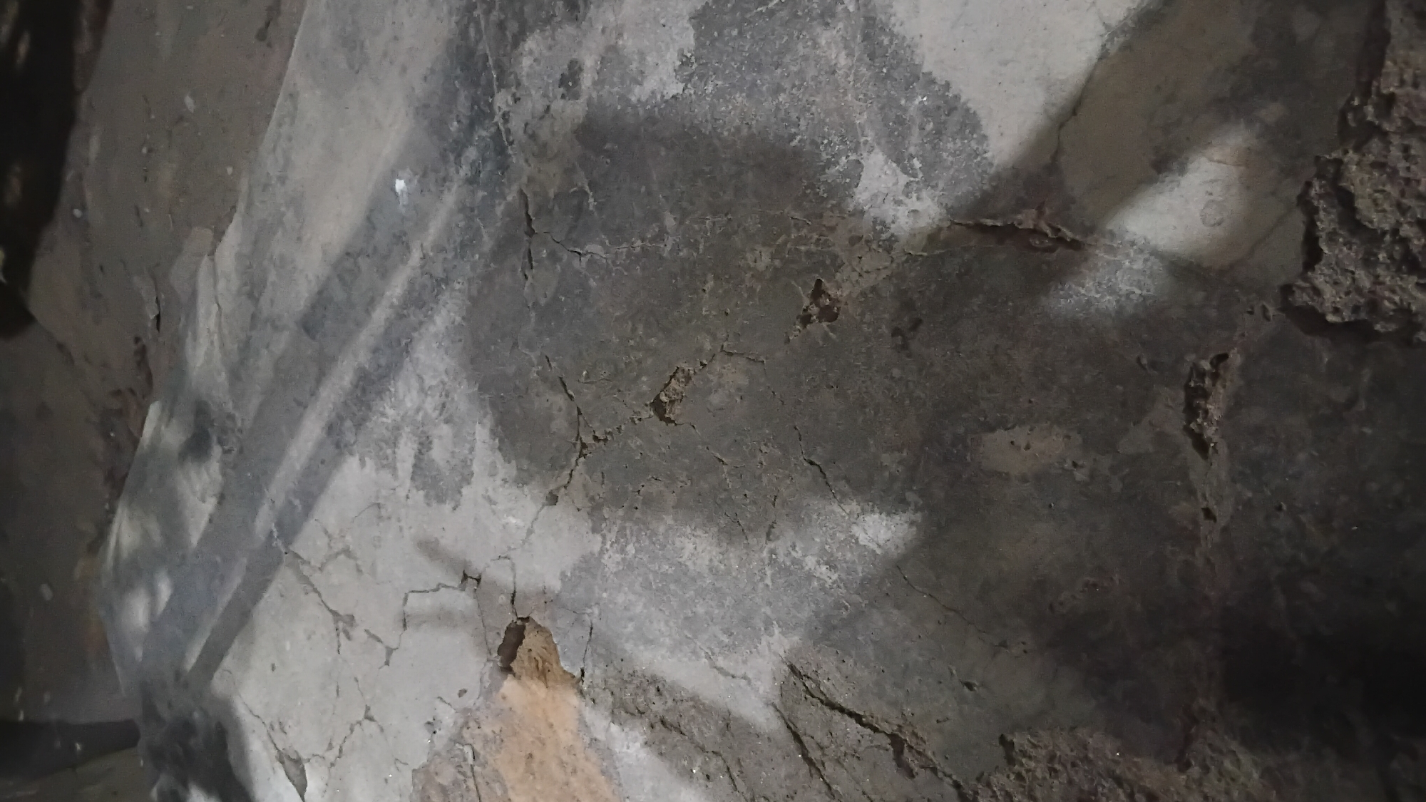 The image depicts a cracked concrete surface, possibly a pathway or a section of ground, in what appears to be an outdoor urban environment. The surface is uneven and shows signs of wear and tear, with multiple cracks and some areas of discoloration or staining. In the background, there are indistinct shadows cast by an unseen light source, suggesting it is either nighttime or the area is heavily shaded. The overall impression is of a neglected or utilitarian urban space. Without further information or context, it is impossible to definitively place this scene in Prabumulih, Indonesia.
