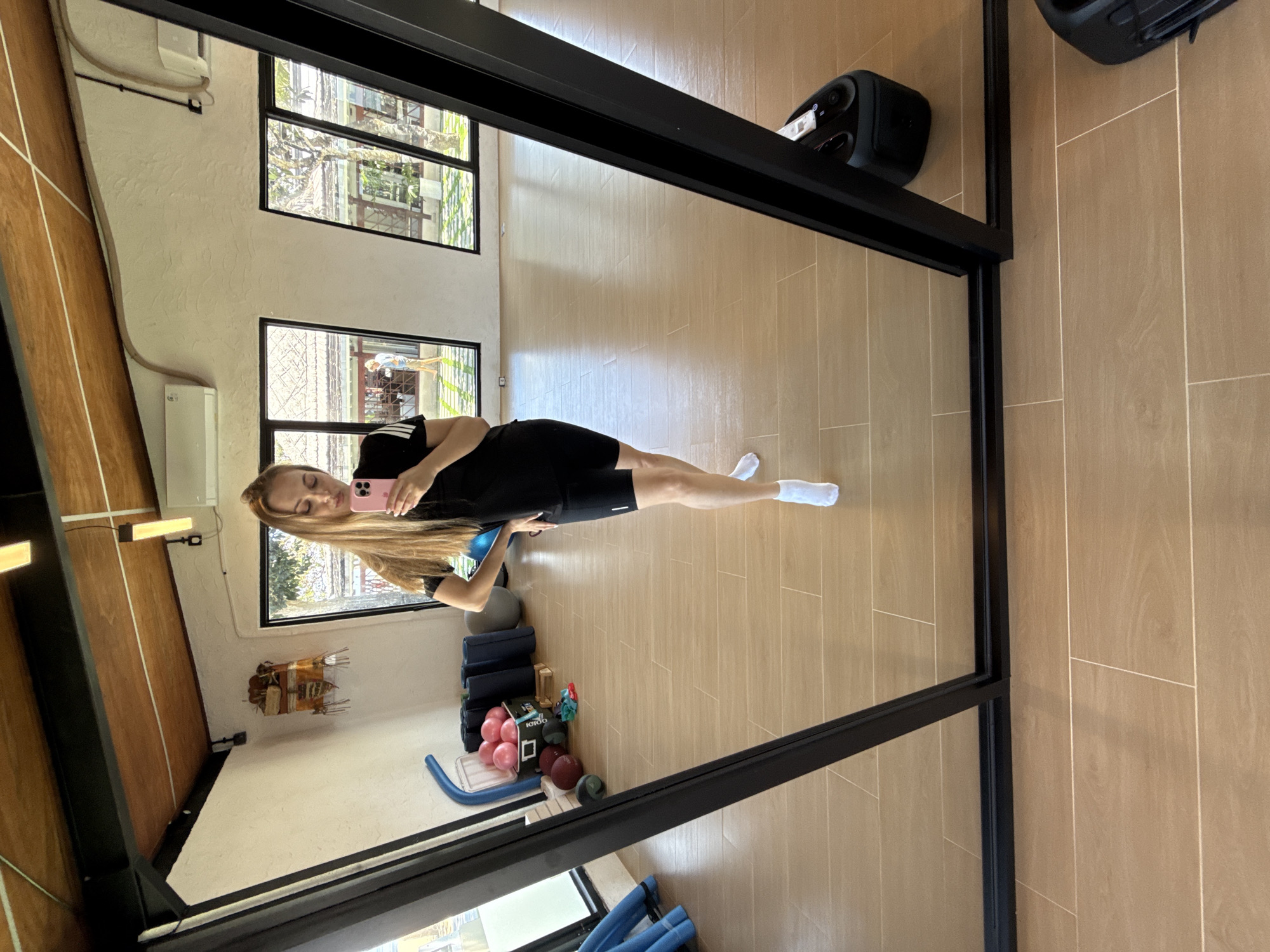 A young woman with long blonde hair, wearing a black athletic top and black shorts with white socks, stands in a gym and takes a selfie in a large mirror. She holds a pink smartphone in her right hand. The gym has light-colored wooden floors and large windows that look out onto a courtyard with trees and traditional Indonesian architecture. In the background, other gym equipment is visible, including yoga mats and balls. The woman appears to be in Denpasar, Indonesia, given the architectural style visible through the windows.