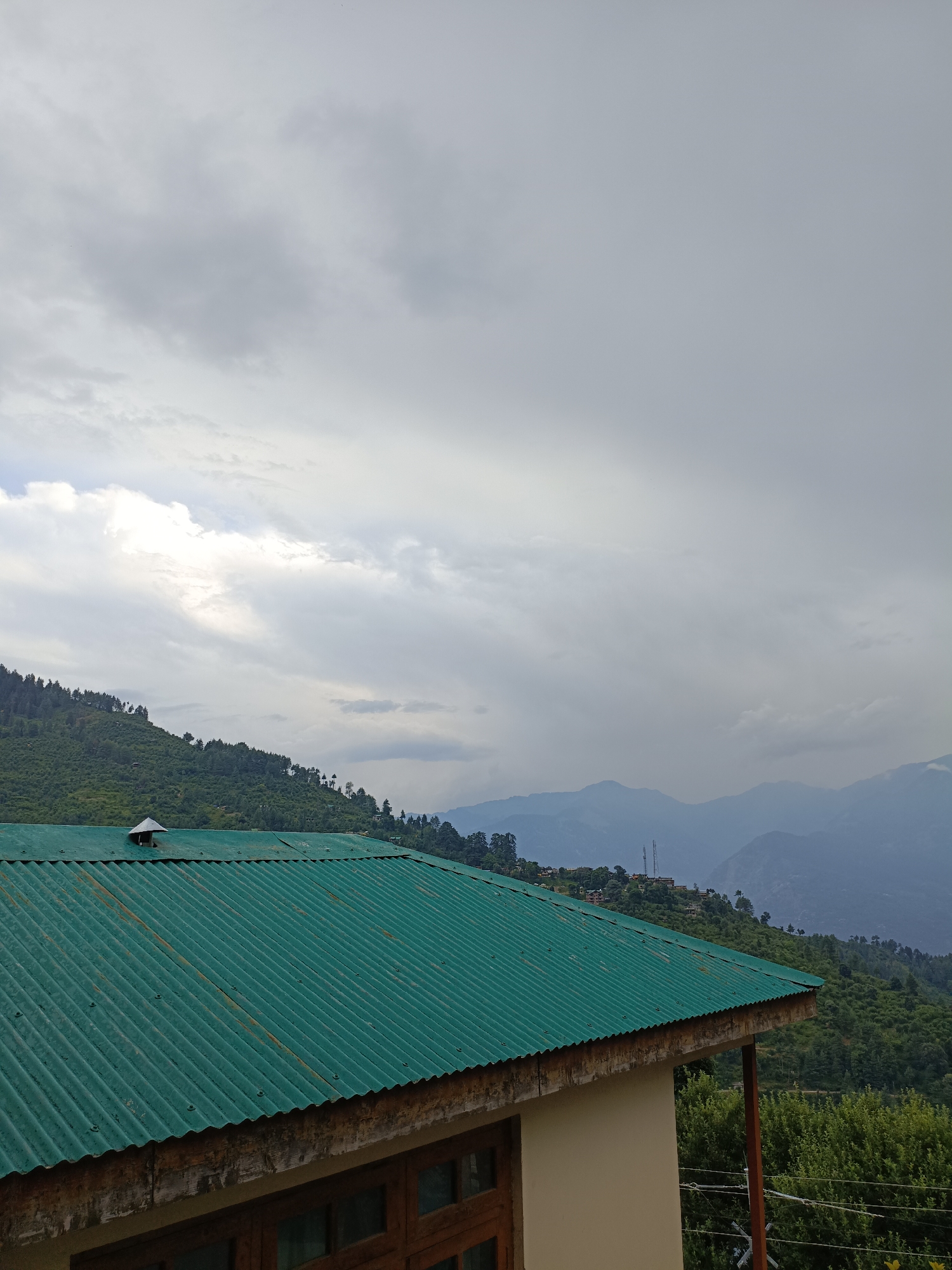 The image depicts a scenic view of a mountainous region, likely in or near Damadi, India, under a cloudy sky. In the foreground, a portion of a building is visible, featuring a corrugated green metal roof with visible screws and some rust spots. The building's exterior wall is painted a light color, and a wooden-framed window is partially visible. A small, pointed vent protrudes from the roof.
Behind the building, a lush green, tree-covered mountainside slopes upwards. The trees are dense, with visible variations in shades of green. The middle ground features more distant mountains, also covered in vegetation, with some scattered structures that suggest a small settlement. In the background, several layers of misty, blue-hued mountains recede into the distance, indicating a vast mountainous landscape. The sky is filled with heavy, grey clouds, with some lighter patches suggesting breaks in the cloud cover or sunlight filtering through. The overall atmosphere is one of natural beauty and a slightly overcast day. No people are present in the image.