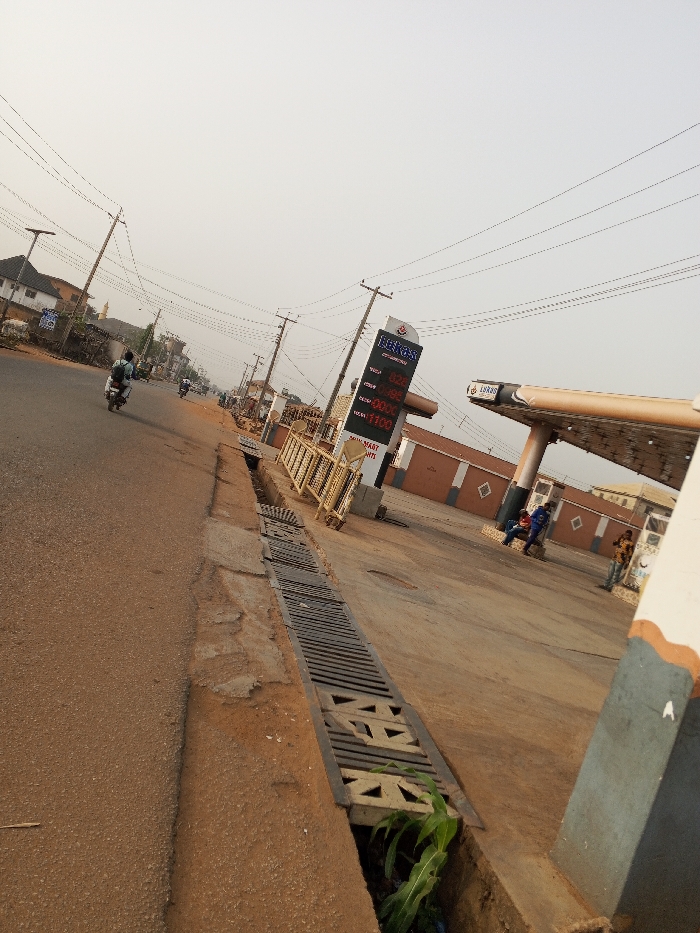 The image captures a slightly angled view of a street scene, dominated by a gas station on the right and a road with a drainage ditch on the left. The perspective is looking down the street, slightly tilted upwards.
On the left side of the frame, a paved road, appearing reddish-brown, stretches into the distance. Alongside the road, a long concrete drainage ditch is visible, covered by metal grates. In the immediate foreground, within the ditch, some green leafy vegetation (possibly corn sprouts) is growing. Utility poles with numerous overhead power lines line the road, receding into the background. A person on a motorcycle is riding away from the camera in the mid-ground on the road, and another motorcycle can be seen further down the street.
The right side of the image features a gas station. A tall, prominent digital price sign displays fuel prices. The brand name "HEKOS OIL & GAS" is visible at the top. Below it, the prices are listed:
* PMS: 828
* AGO: 790
* DPK: XXX (partially obscured, but the last digit looks like 0)
* LUSSI: 1100
Below these prices, smaller text includes "LUCKY SMART POINTS" and what appears to be phone numbers.
Adjacent to the sign is a gas station canopy structure, supported by pillars, with the "HEKOS" brand logo also visible on its edge. Behind the canopy, there are low-rise, single-story buildings painted brown with white horizontal trim. Several individuals are present at the gas station; two men are sitting on a low wall or bench near the buildings, and another person is standing further to the right.
The sky is a pale, hazy off-white or light blue, suggesting either a clear but somewhat dusty or humid day, or perhaps early morning/late afternoon with a muted light. The overall lighting is bright and even, with soft shadows. The general setting appears to be a typical urban or semi-urban environment.