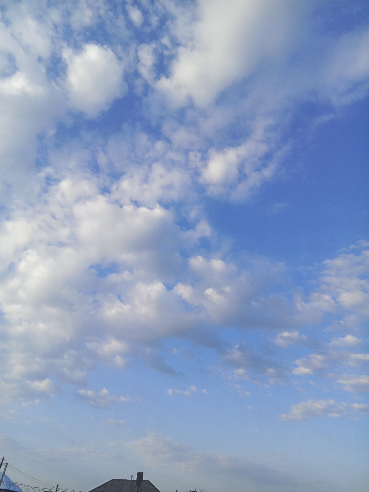 The image shows a bright blue sky with scattered white cumulus clouds. The clouds are varied in size and density, with some appearing wispy and others more substantial. The lower portion of the image reveals a sliver of the tops of buildings and what appears to be a barbed wire fence, suggesting an urban or suburban environment. The overall impression is of a clear and pleasant day.