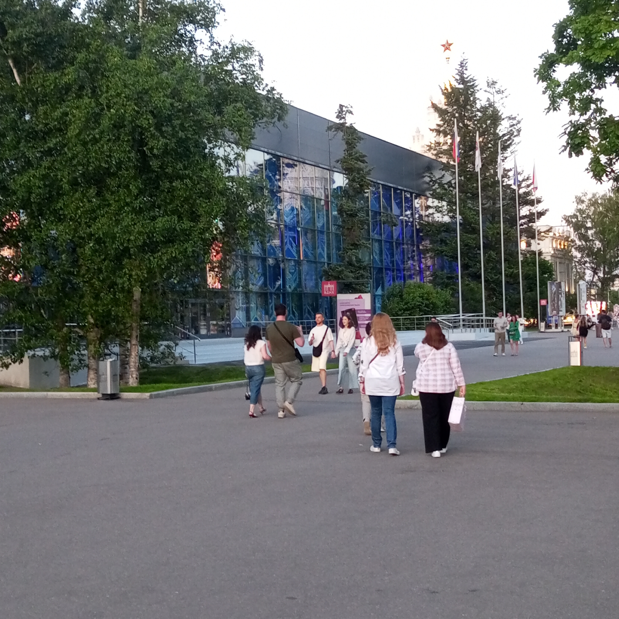This image captures an outdoor scene at VDNKh (Exhibition of Achievements of National Economy) in Moscow, Russia, during daylight hours.
In the foreground, a wide, dark gray asphalt path occupies the lower half of the frame. On the path, six individuals are walking away from the viewer. From left to right: a woman in a white top and blue jeans with a black bag; a man in a dark green t-shirt and light pants with a black messenger bag, looking towards the woman; a woman with light brown hair in a white shirt and blue jeans with a white shoulder bag; and a woman in a pink and white checkered shirt and black pants holding a light-colored rectangular object. Two additional individuals are partially visible behind the main group.
To the left of the path, dense green foliage from large trees covers a significant portion of the midground and extends into the upper left. A tall, cylindrical, metallic object is positioned near the path's edge amidst the trees. To the right of the path, a strip of green grass borders the asphalt.
In the midground, a large building with a modern facade dominates the background. This building features numerous dark blue reflective glass panels, displaying reflections of trees and other architectural elements. The upper section of the building is dark gray. A small red sign with white text, possibly "ВДНХ," is affixed to the glass facade.
To the right of this building, several tall, white flagpoles stand, two of which display flags: one is the Russian tricolor (white, blue, red), and the other is a white or light-colored flag. Beyond the flagpoles and trees, a light-colored, monumental building with a prominent red five-pointed star on its spire is visible against the bright, overcast sky. This structure is characteristic of the Central Pavilion or Main Entrance Arch at VDNKh.
Further in the background, more indistinct figures are scattered across paved areas, engaged in walking and conversing. The overall environment suggests a well-maintained public space or exhibition ground.