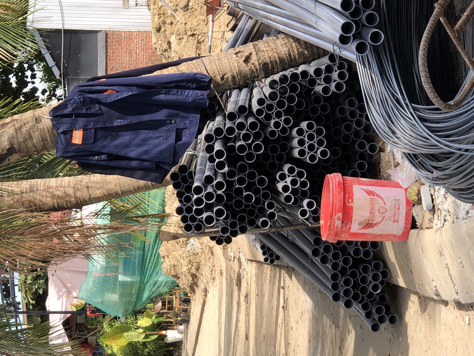 The image depicts a construction site in Đà Nẵng, Vietnam, with a focus on materials and workwear. A dark blue jumpsuit with orange reflective strips is hanging on a palm tree trunk. Next to it, a large pile of grey plastic pipes is stacked, with a red bucket labeled "SINGAPORE" positioned in front of them. To the right, a bundle of coiled metal rebar is visible, along with more grey pipes. In the background, there is evidence of ongoing construction, including piles of dirt, a brick wall, and a corrugated metal building. The scene suggests a temporary outdoor workspace.