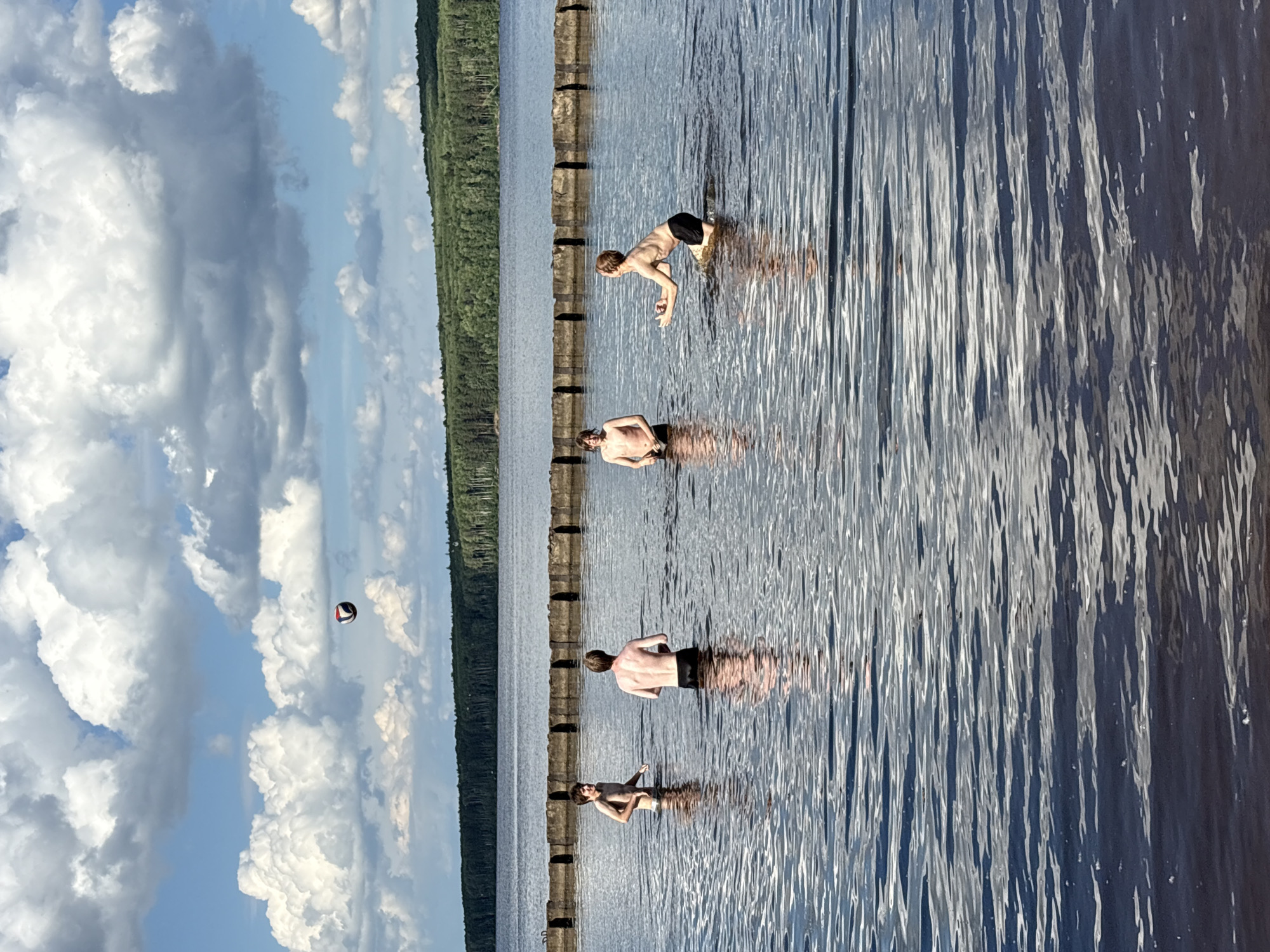 In a wide body of water, likely a lake or reservoir, four young males are engaged in a game of volleyball. The water is calm with gentle ripples, reflecting the bright sky and white, puffy clouds. The young men are partially submerged, with water reaching to their waists. They are dressed in swimwear. One individual in the foreground right is in the process of catching or hitting the volleyball, which is suspended in the air above and slightly to his left. Another individual, to his left and further back, is also looking at the ball. Two other individuals are visible further to the left. In the background, a line of what appears to be a man-made barrier, possibly a breakwater or a series of pilings, stretches across the water. Beyond the water, a dense, green forest lines the horizon under a partly cloudy sky. The scene suggests a recreational activity on a warm, sunny day. The specific location in Chaikovsky, Russia cannot be determined from the image alone.