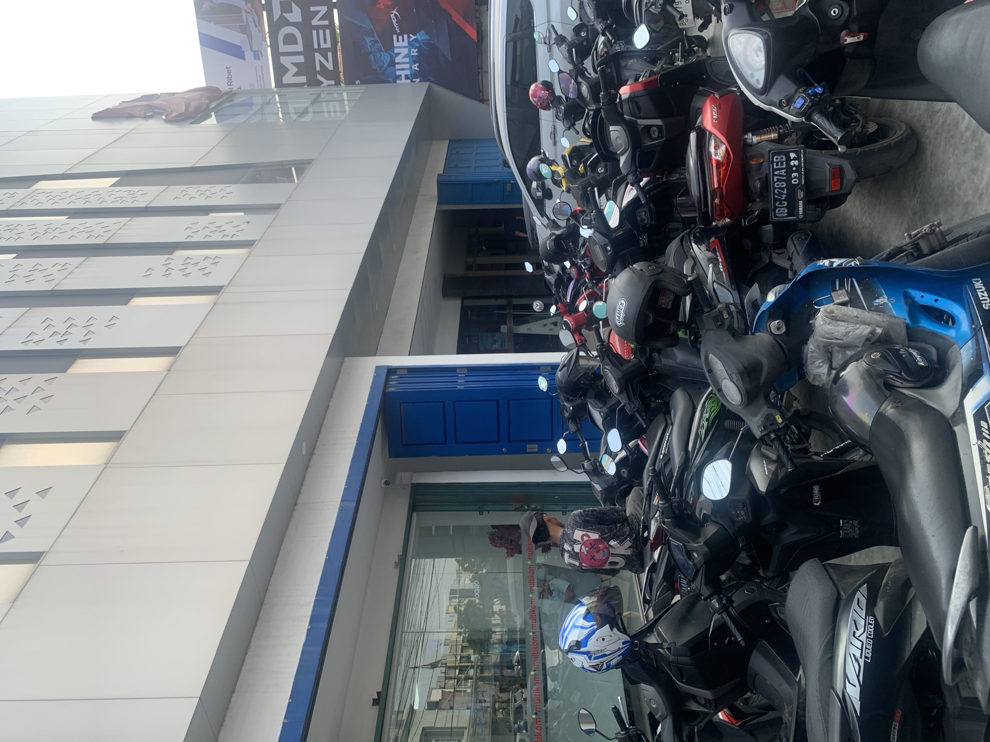 The image shows a street scene in Palembang, Indonesia, likely in front of a commercial building. The foreground is filled with a large number of parked motorcycles, some with helmets on them. A white car is visible behind the motorcycles, partially obscured. The building has a modern facade with white paneling and decorative triangular cutouts. A large banner on the building advertises "AMD RYZEN" and "MACHINE". A person wearing a dark t-shirt with a pink graphic and a cap is sitting on one of the motorcycles, facing away from the camera. Another person, wearing a patterned shirt and a cap, is visible through the storefront window. The storefront appears to be a retail establishment, with products visible inside. The overall impression is a busy urban environment with a focus on retail and transportation.