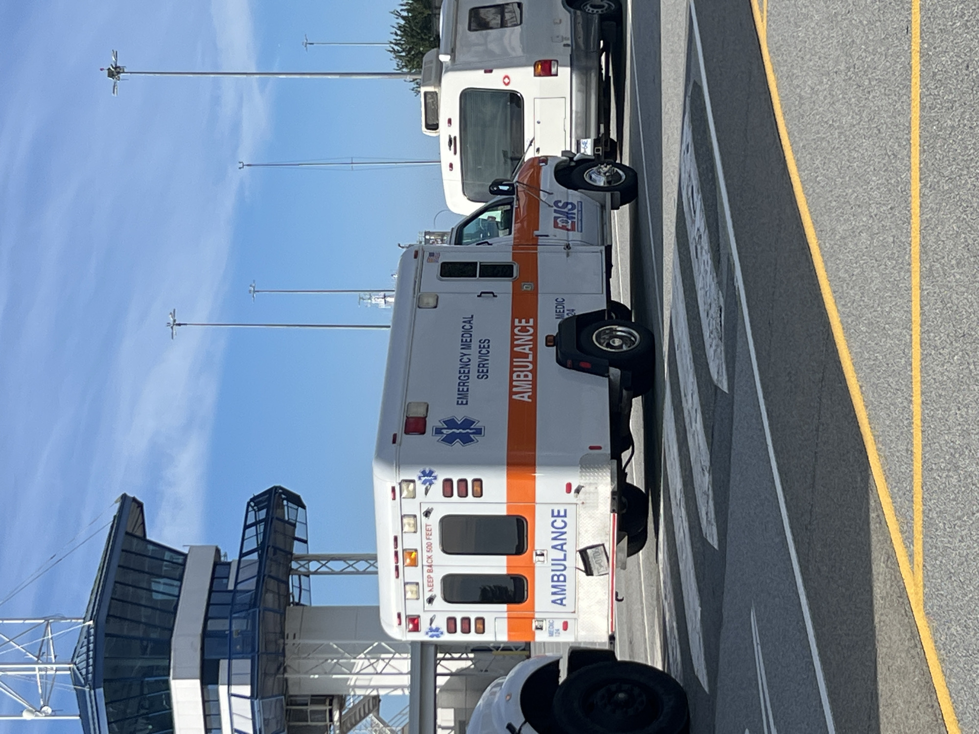 An exterior, medium shot captures an ambulance and a white camper van parked on an asphalt road under a clear blue sky. The ambulance, predominantly white with orange accents, has "AMBULANCE" written in blue on its rear. A blue Star of Life emblem and "EMERGENCY MEDICAL SERVICES" are visible on its side. The camper van is parked to the right of the ambulance. In the background, a tall, modern control tower with glass windows stands against the sky, accompanied by tall light poles. The scene appears to be at an airport or a large transportation hub. The location is identified as the city of Delta, Canada.