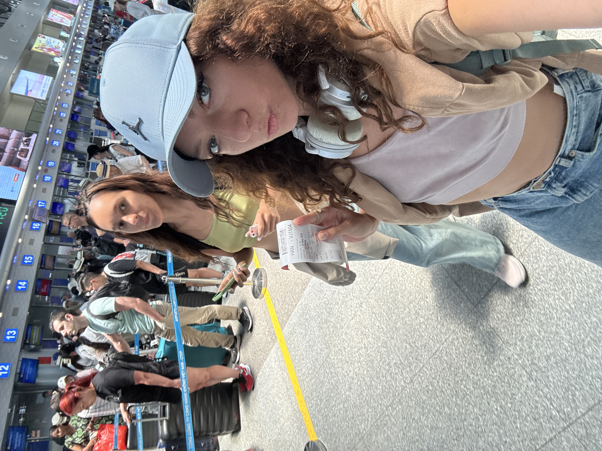 The image shows two women at the airport. The woman in the foreground is wearing a baseball cap, tank top, and jeans with headphones around her neck. She's holding up a boarding pass. The other woman is standing slightly behind her. Behind them are lines of people waiting at check-in counters, with digital displays visible above the counters. One boarding pass is visible. It says "HAN-VJ1504 Hanh Khach Passenger ANGELOVA ALISA. 15 May 2026 De Nang"