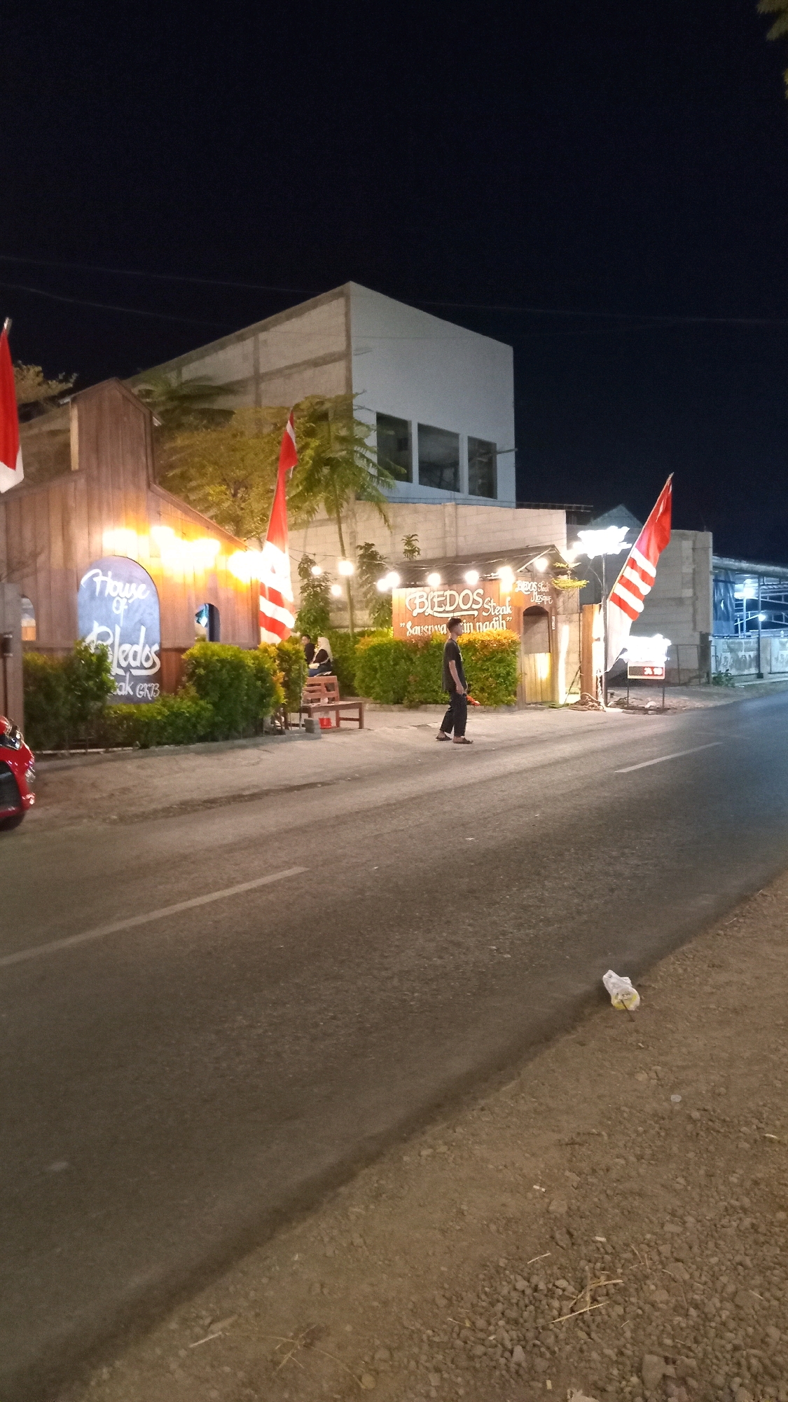 This is a nighttime street-level photograph taken in front of a restaurant in Suci, Indonesia. The establishment appears to be called "House of Bledos Steak". A red car is partially visible on the left side of the image. In front of the restaurant, a man in dark clothing walks on the sidewalk, and two individuals, one seated on a bench, are visible near the entrance. Red and white Indonesian flags are displayed on poles. The street in the foreground is paved, with gravel and debris along the right side. The background shows a multi-story building and other structures. String lights are illuminating the restaurant's facade.