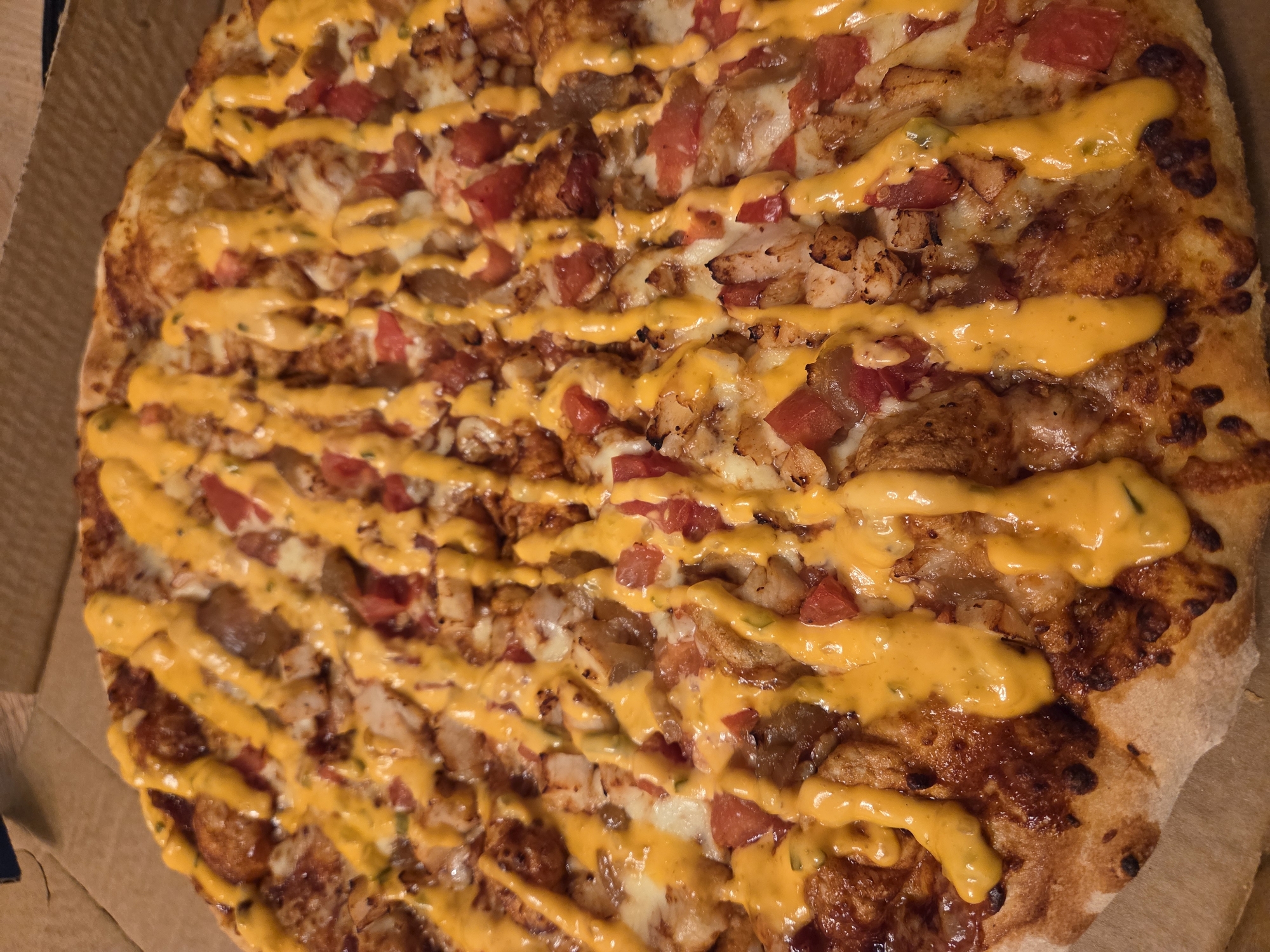 An overhead shot reveals a pizza in a cardboard box. The pizza is topped with what appears to be cheese, diced tomatoes, chicken, and a yellow sauce drizzled in lines across the top. The box material is a light brown color. The pizza is roughly circular and fills much of the box's area. The toppings appear to be evenly distributed across the pizza. There is no evidence to determine that this image was taken in Varna, Bulgaria.