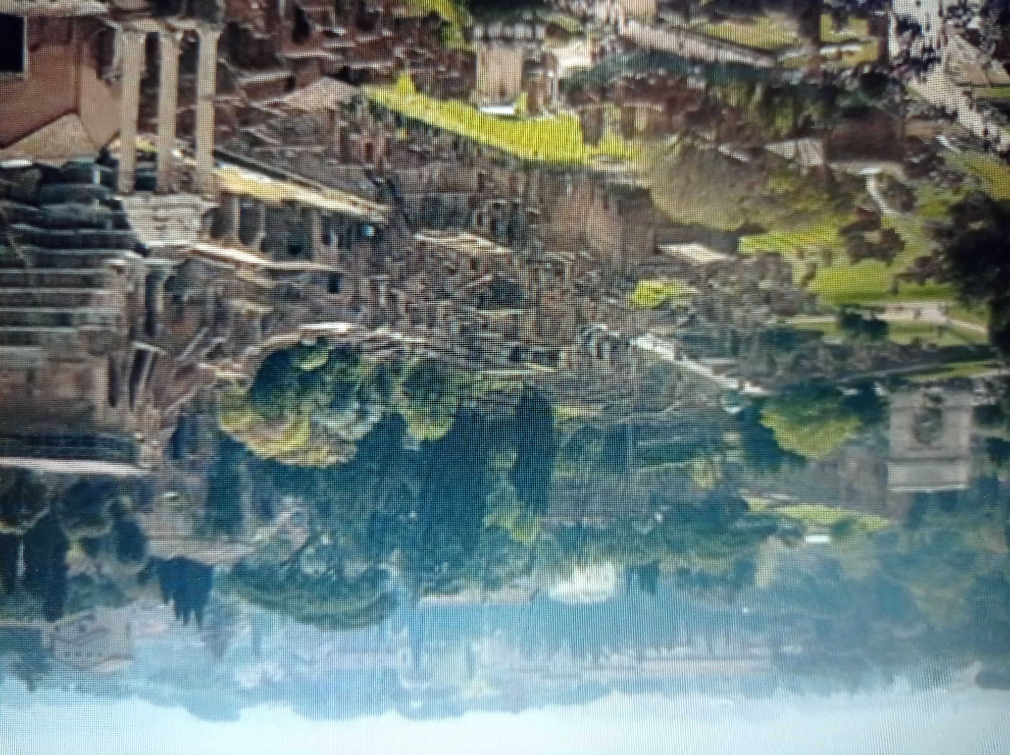 This is a wide-angle, high-angle aerial view of the Roman Forum in Rome, Italy. The scene is dominated by the ruins of ancient Roman structures, including arches, temples, and crumbling walls, interspersed with lush green trees and grass. In the foreground and mid-ground, these ruins form a complex, layered landscape. Several pathways and roads are visible, with a number of people walking along them, particularly on the left side of the image. In the background, more distant buildings and trees form a hazy cityscape, indicating the modern city of Rome surrounding the ancient site. The lighting suggests it is a bright, possibly sunny day, with a slight haze or atmospheric perspective obscuring the furthest details. The image does not depict the city of Mukachevo, Ukraine.